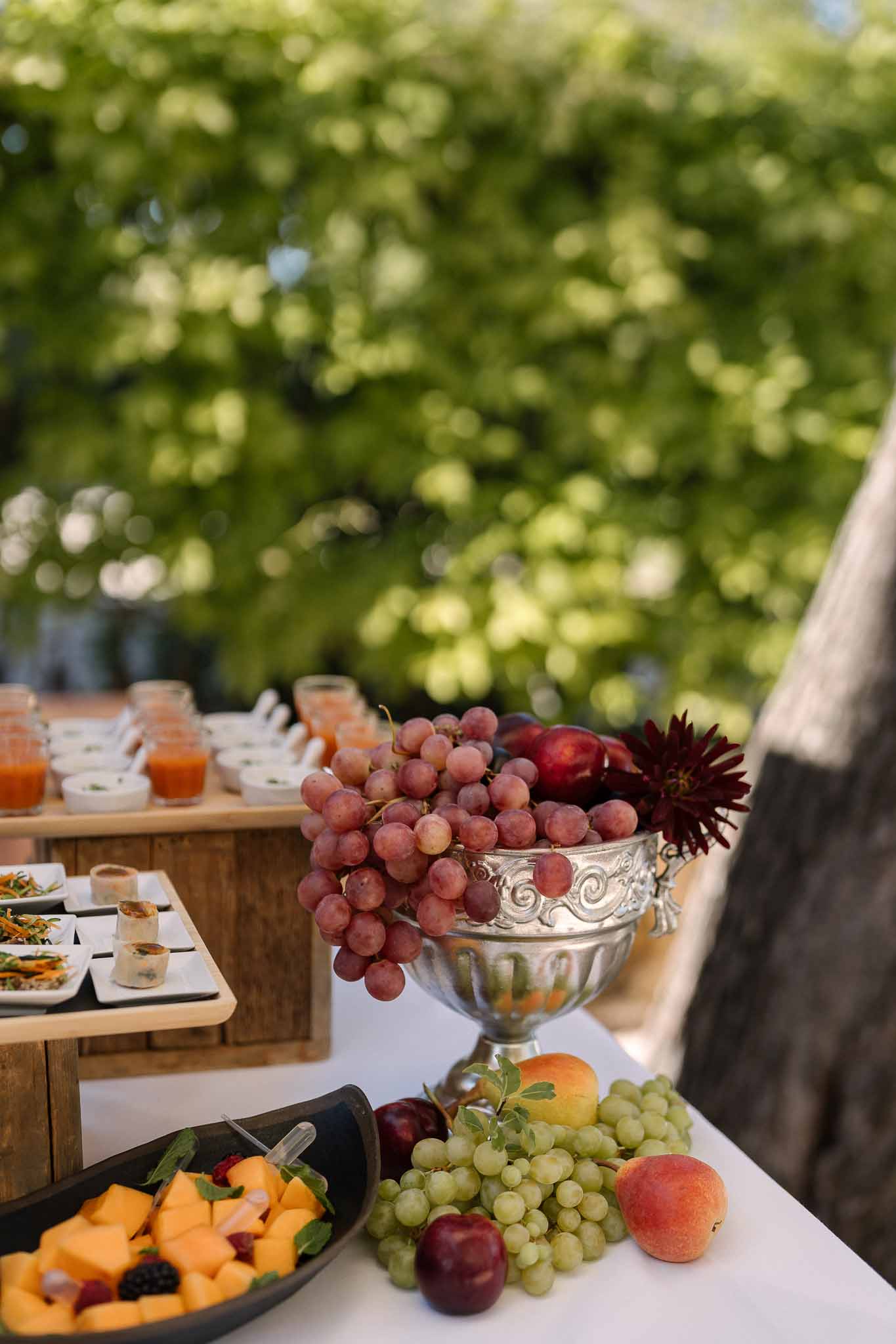 Silver pedestal bowl with grapes, peaches, and burgundy dahlias beside tiered canape display at cocktail buffet
