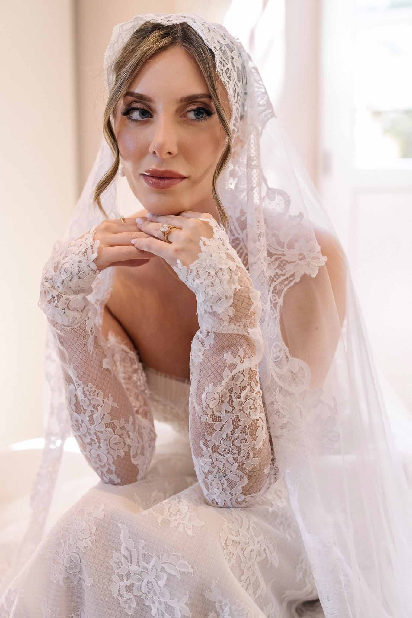 Close-up bridal portrait in lace long-sleeve dress and mantilla veil with hands folded showing oval engagement ring