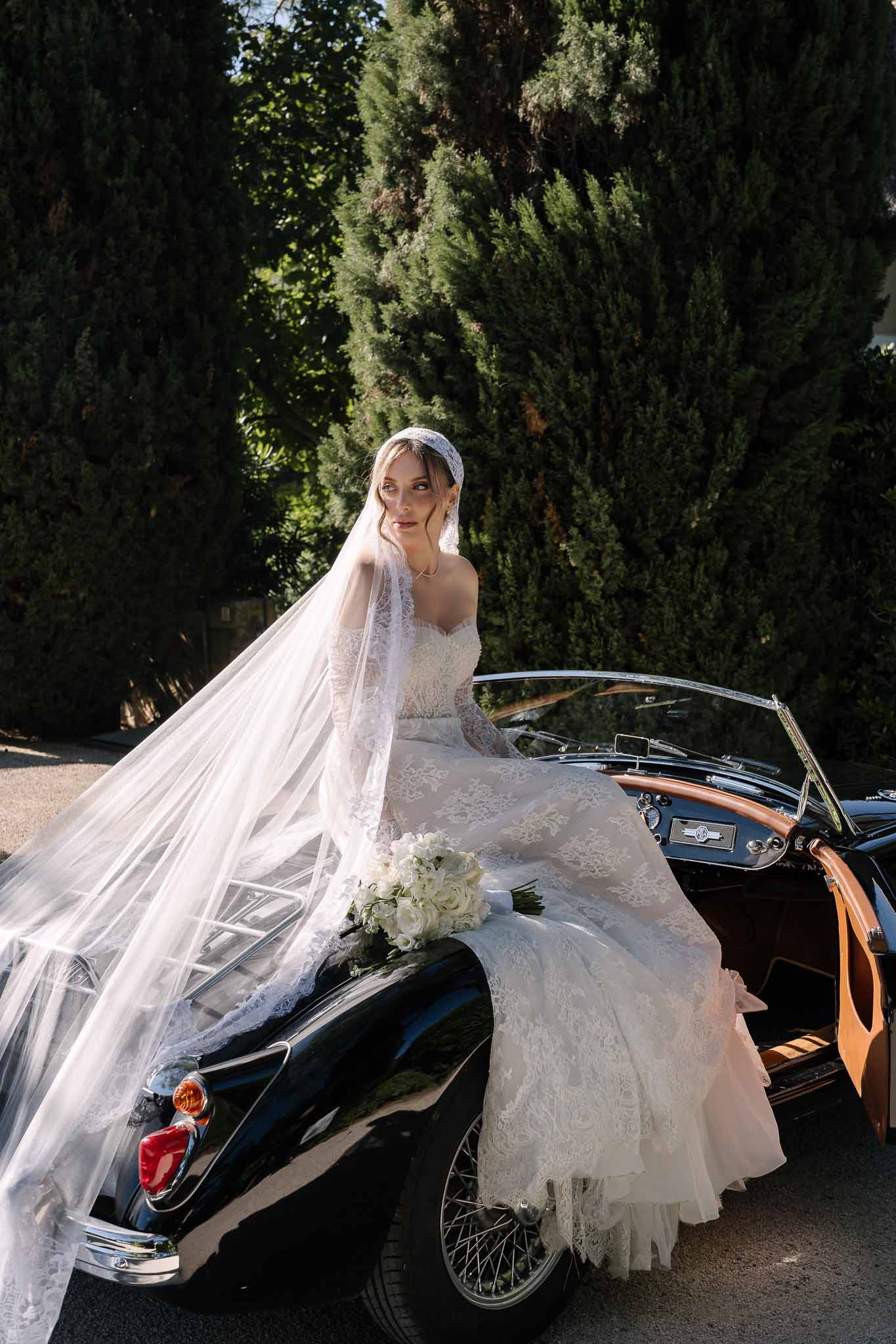 Bride in ivory lace off-shoulder gown and Juliet cap veil seated on vintage black convertible holding white rose bouquet