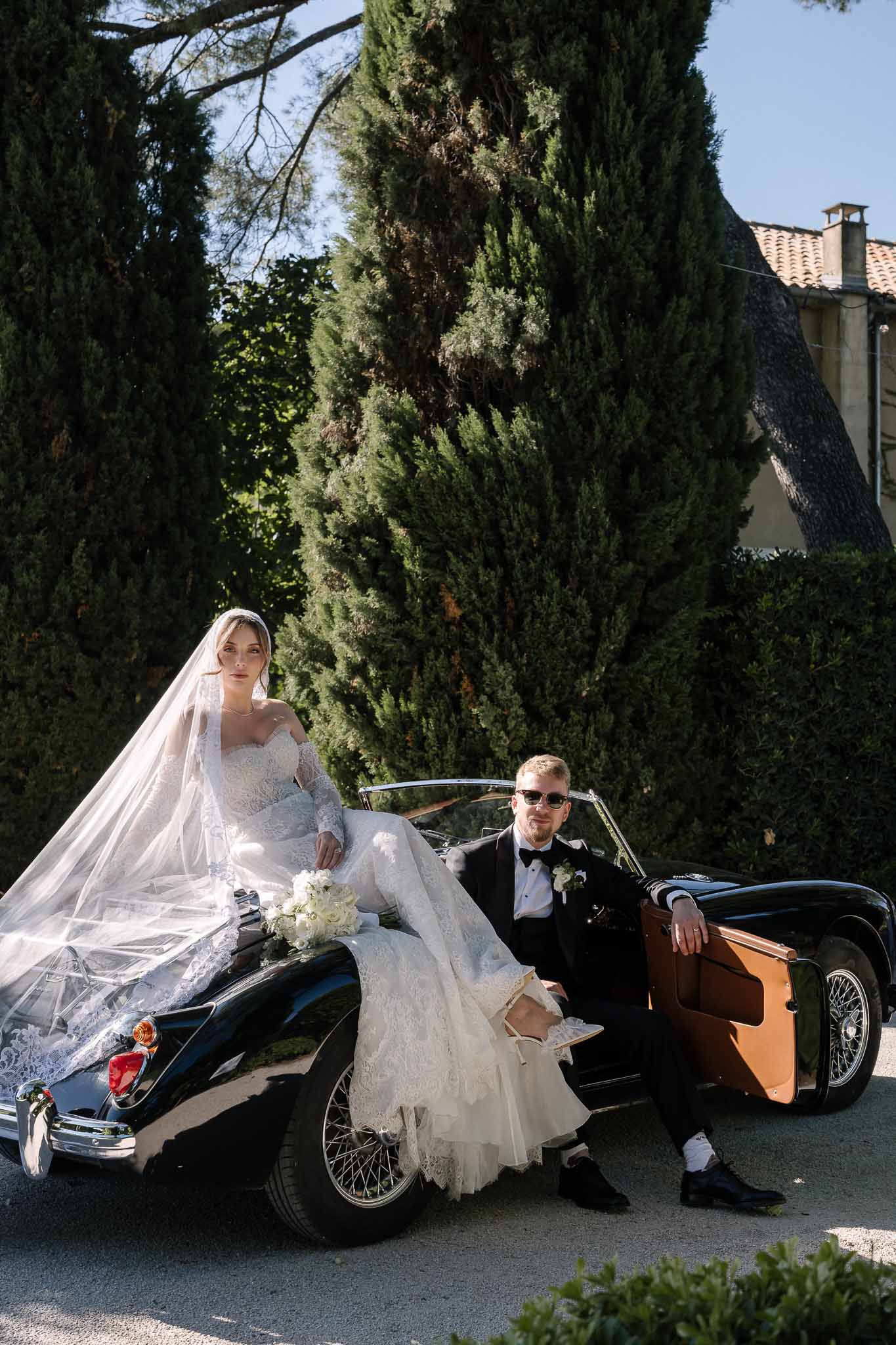 Bride in lace gown with cathedral veil on vintage black convertible beside groom in tuxedo and sunglasses