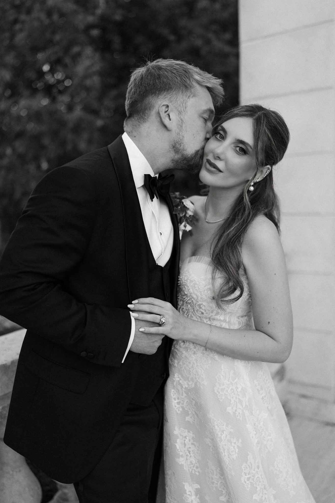 Black and white groom kissing bride's temple with pearl earrings and strapless lace gown against column