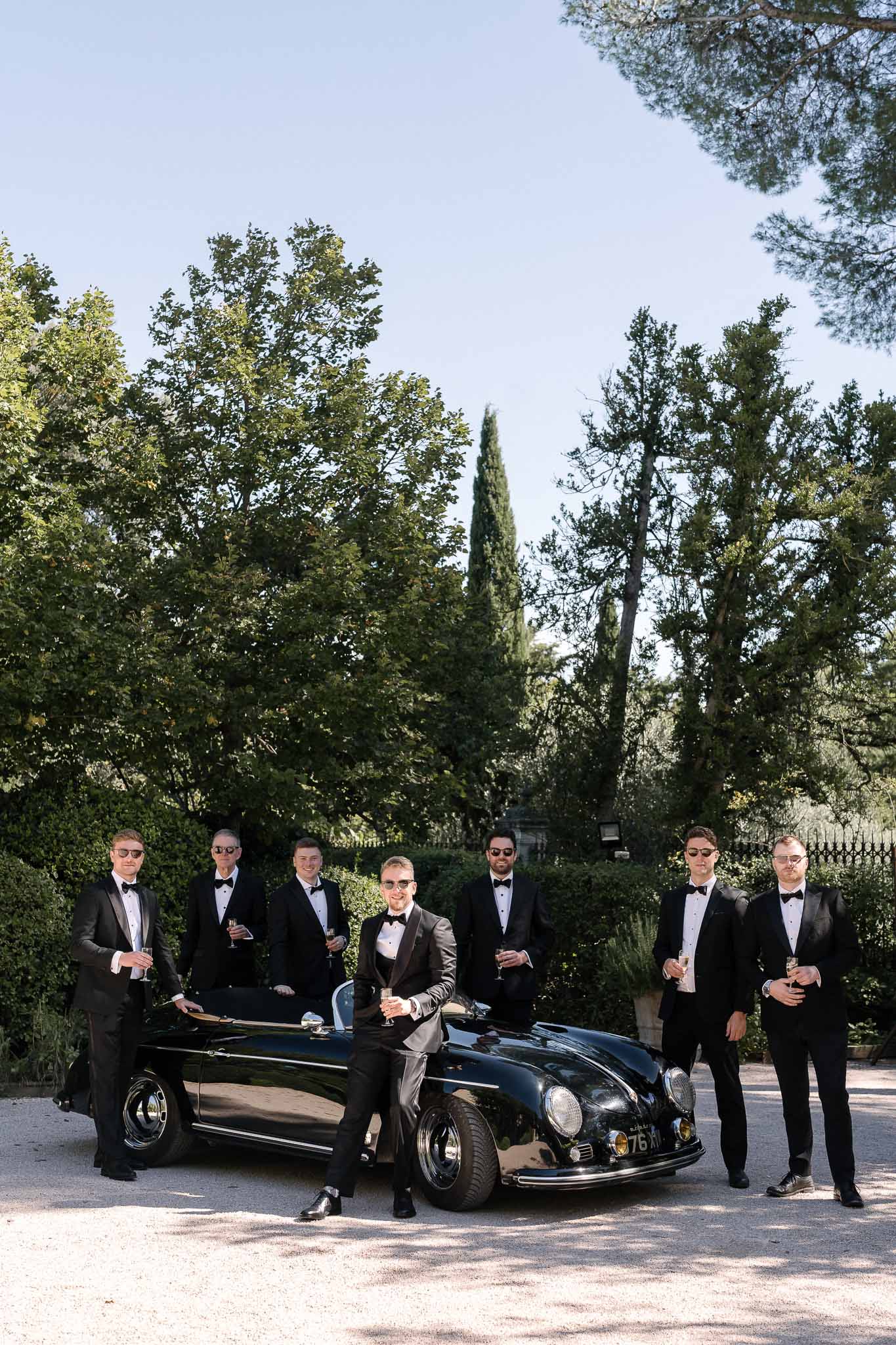 Groom and six groomsmen in black tuxedos with sunglasses pose beside vintage black Porsche convertible