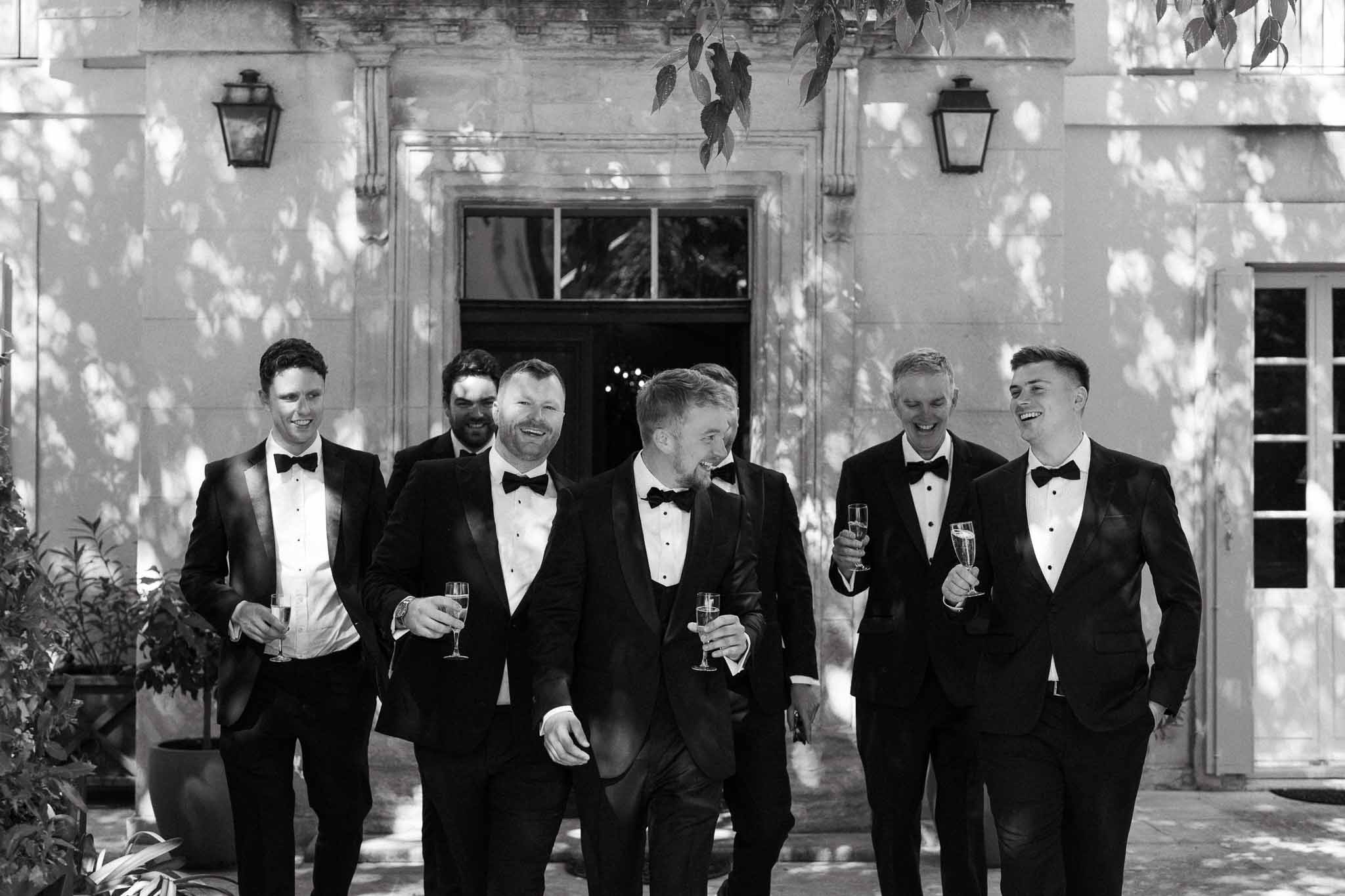 Black and white groom and five groomsmen in tuxedos walking with champagne flutes before chateau with dappled light
