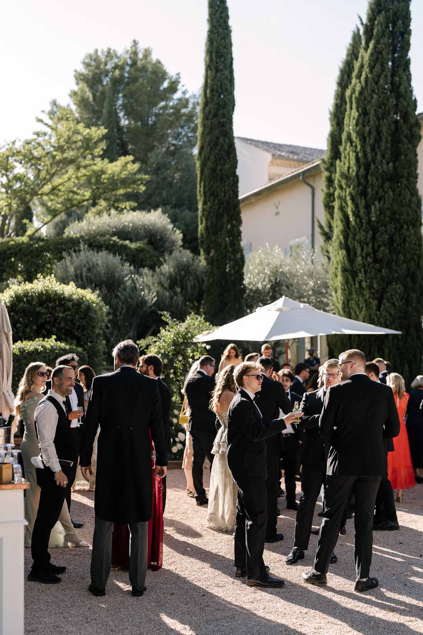 Guests mingling on gravel terrace during cocktail hour at Provencal domaine with cypress trees