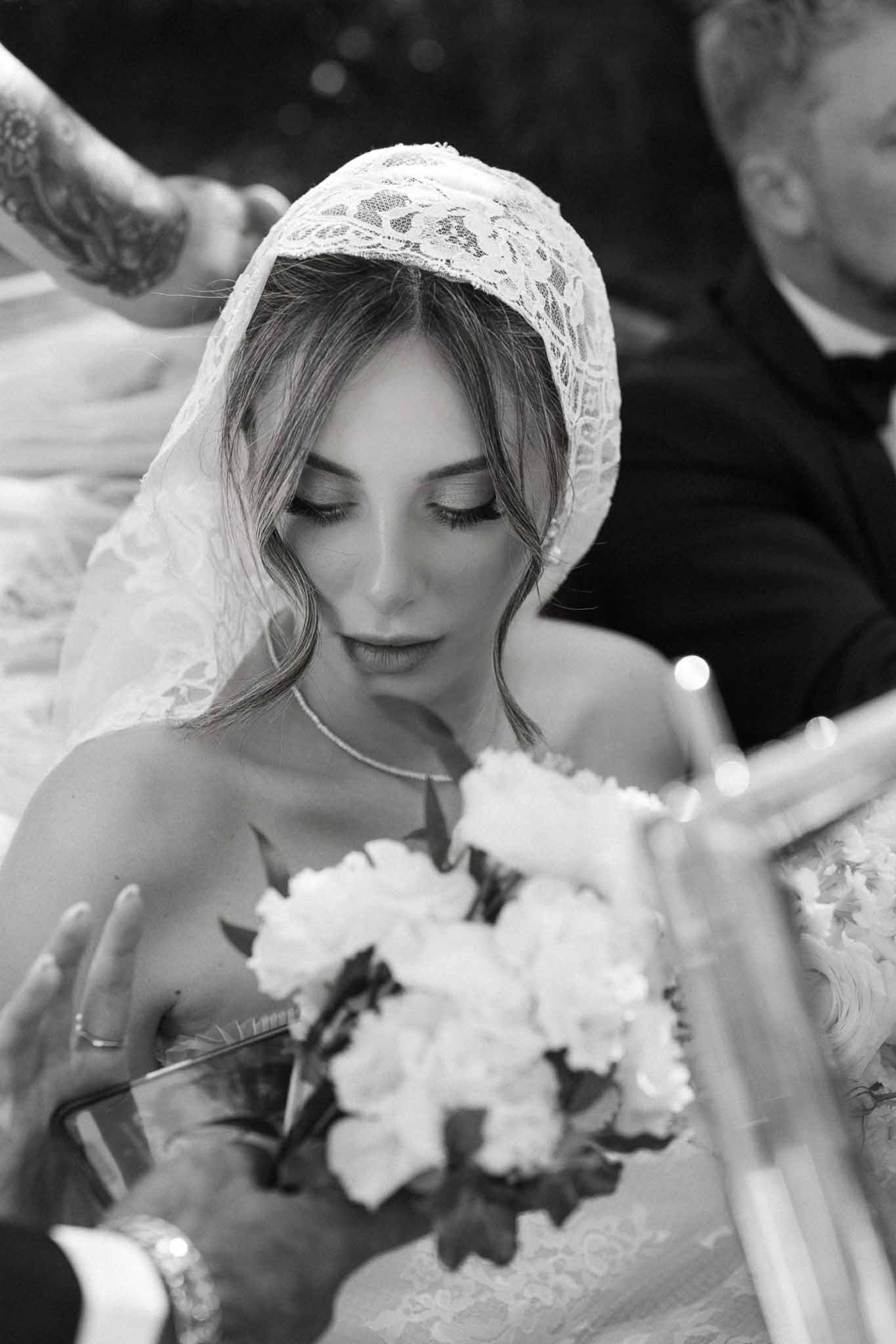 Black and white close-up of bride in lace juliet cap veil holding peony bouquet during ceremony