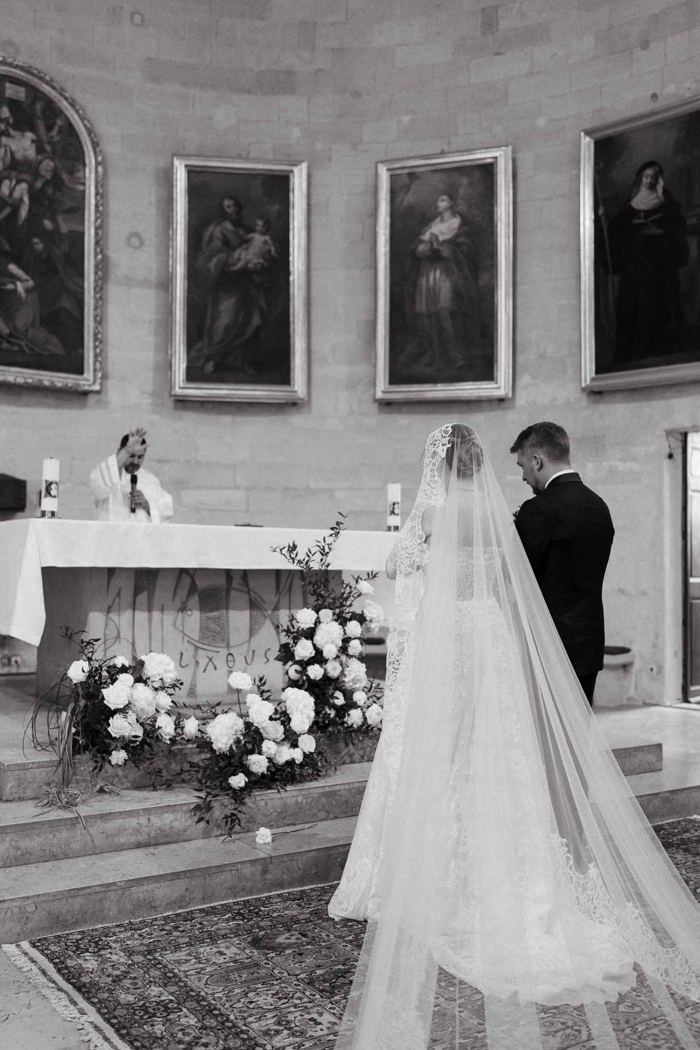 Black and white view from behind couple at church altar with priest and cathedral-length veil