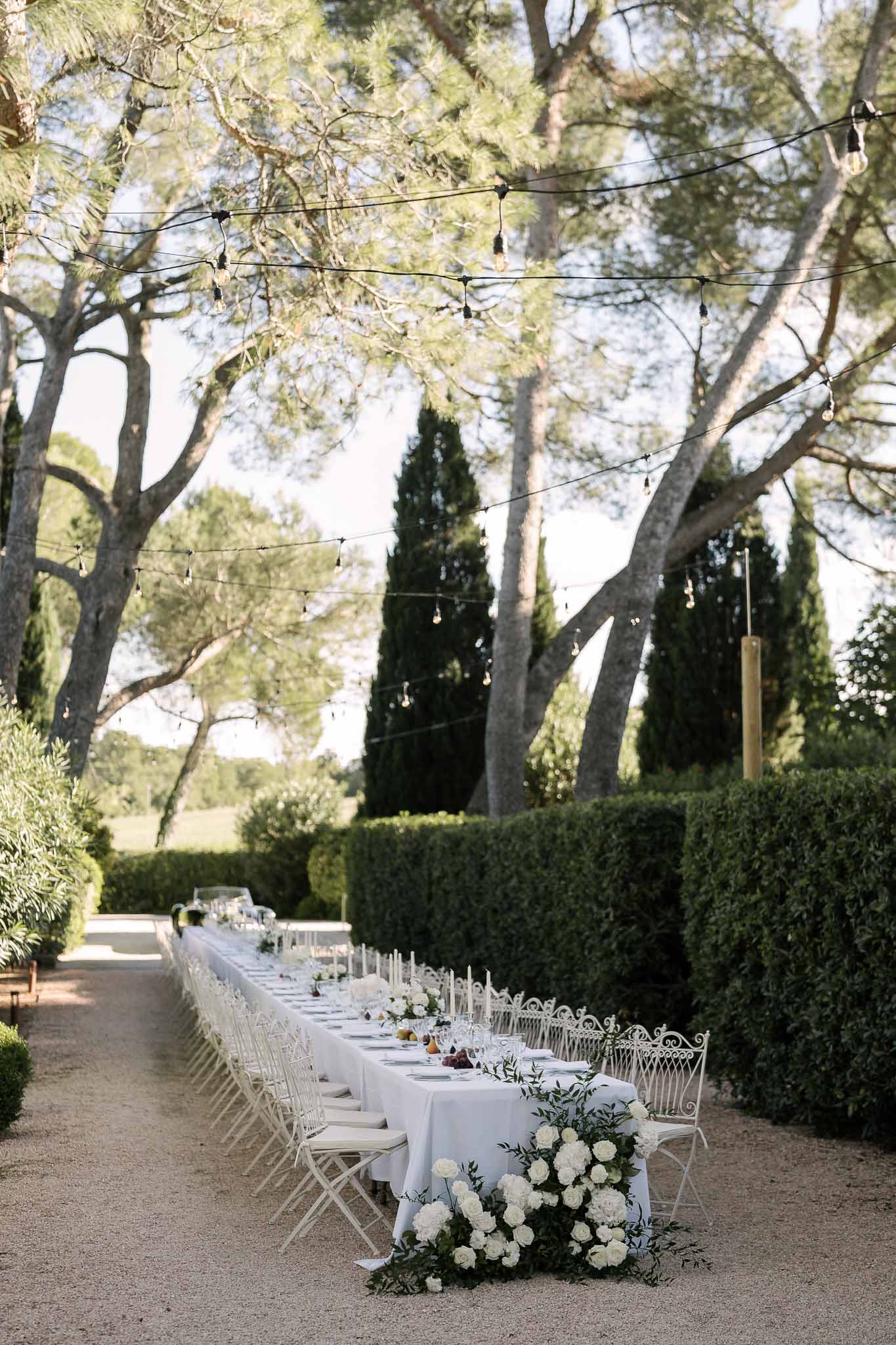 Garden feasting table with white peonies taper candles wrought iron chairs and Edison string lights overhead
