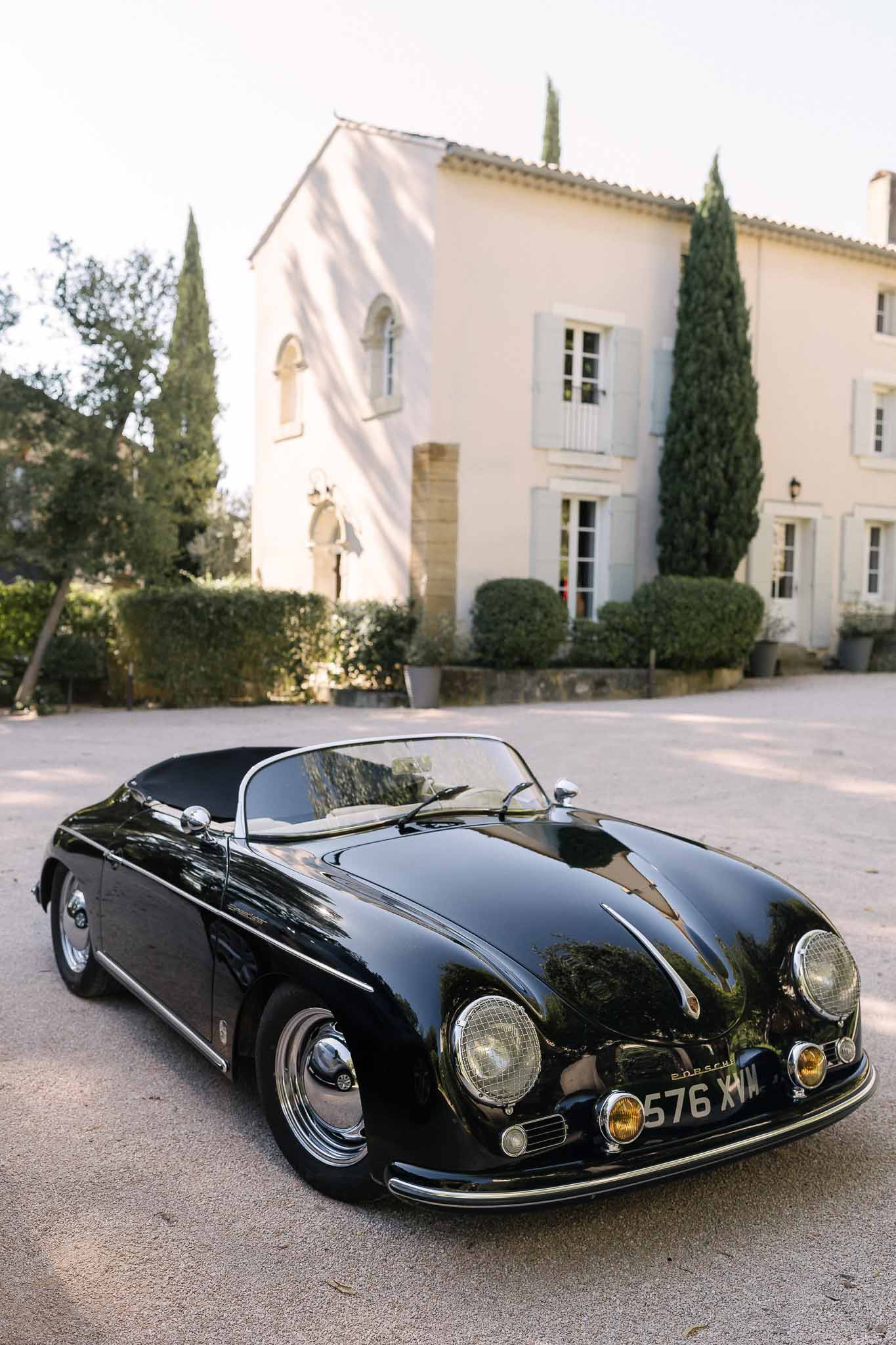 Black vintage Porsche 356 Speedster on gravel courtyard before cream Provencal bastide with blue shutters and cypress trees