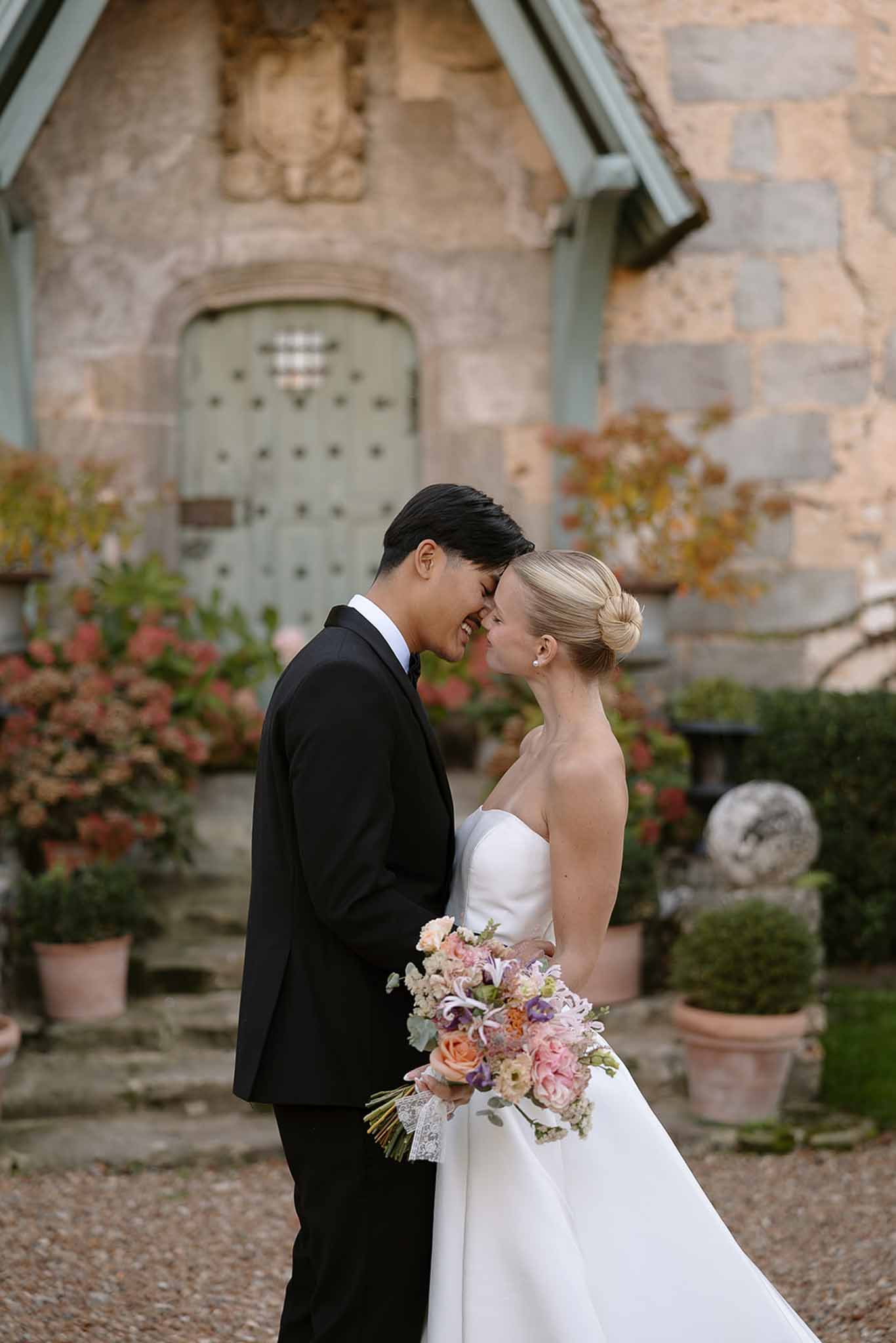 Couple forehead-to-forehead at sage-green chateau door, bride with peach and lavender bouquet