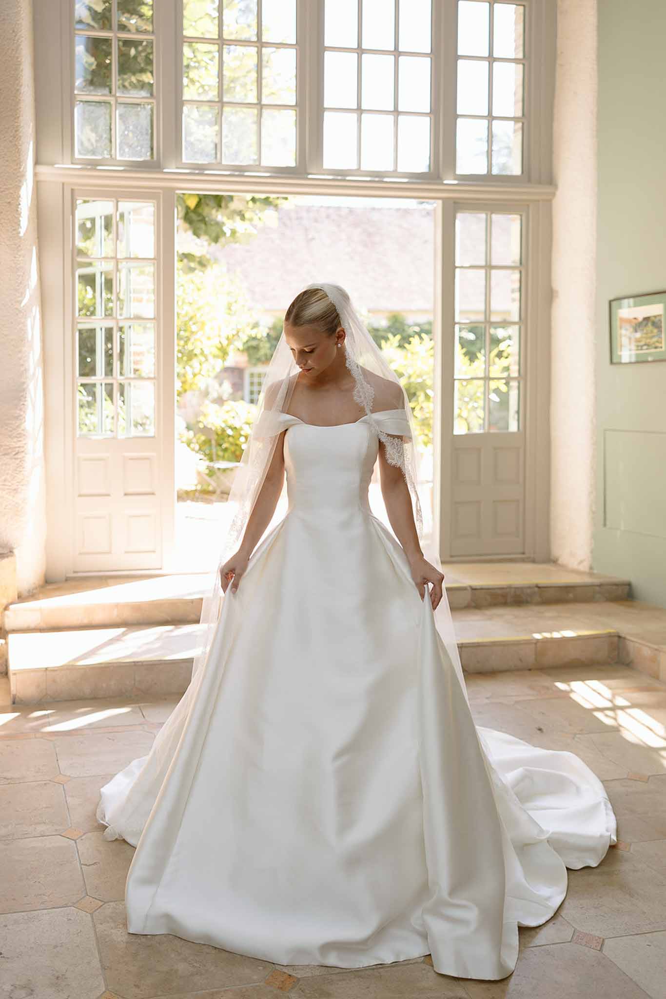 Bride in off-the-shoulder white ballgown and cathedral-length veil stands by French doors in chateau interior