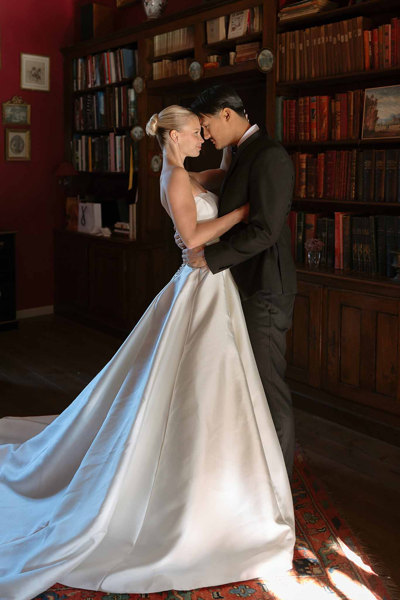 Couple embracing with foreheads touching in deep red library with floor-to-ceiling leather-bound bookshelves