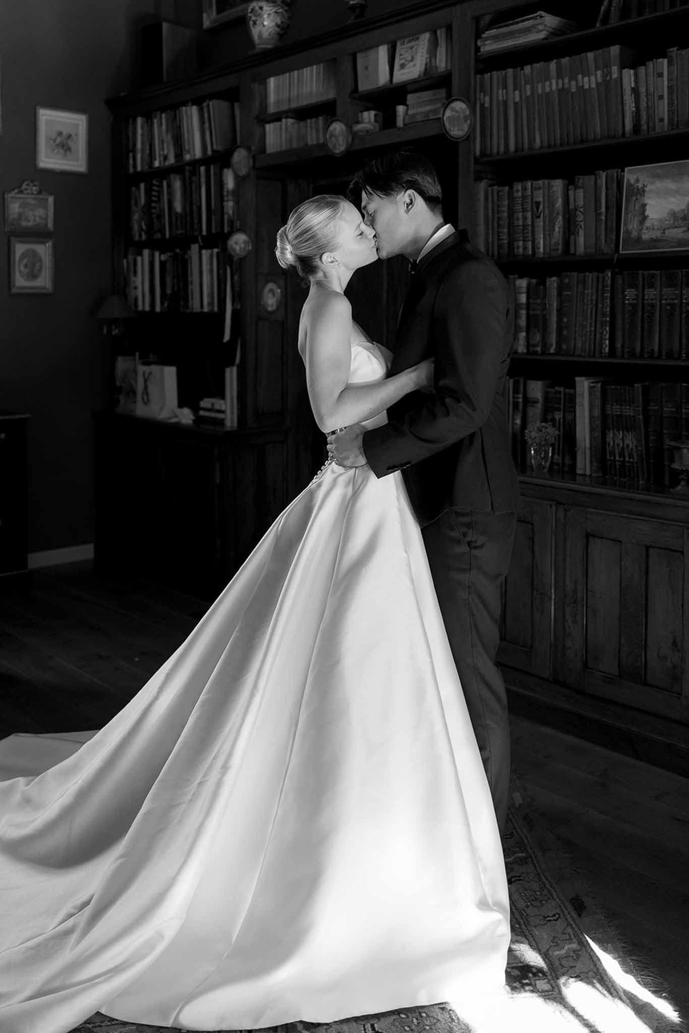 Black and white photo of bride and groom kissing in wood-panelled library with dress train on parquet floor