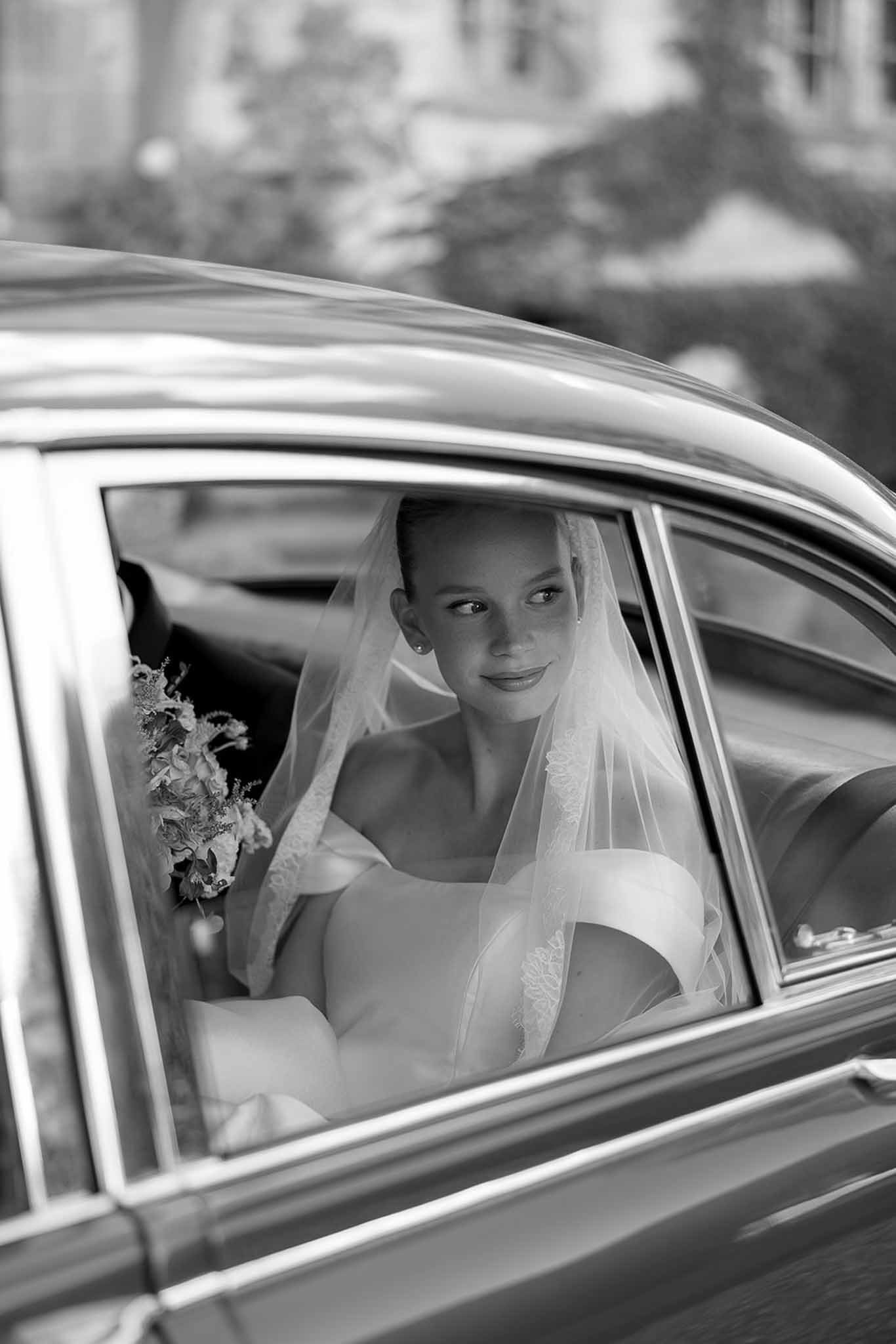 Black and white portrait of bride holding bouquet through classic car window with lace-trimmed veil