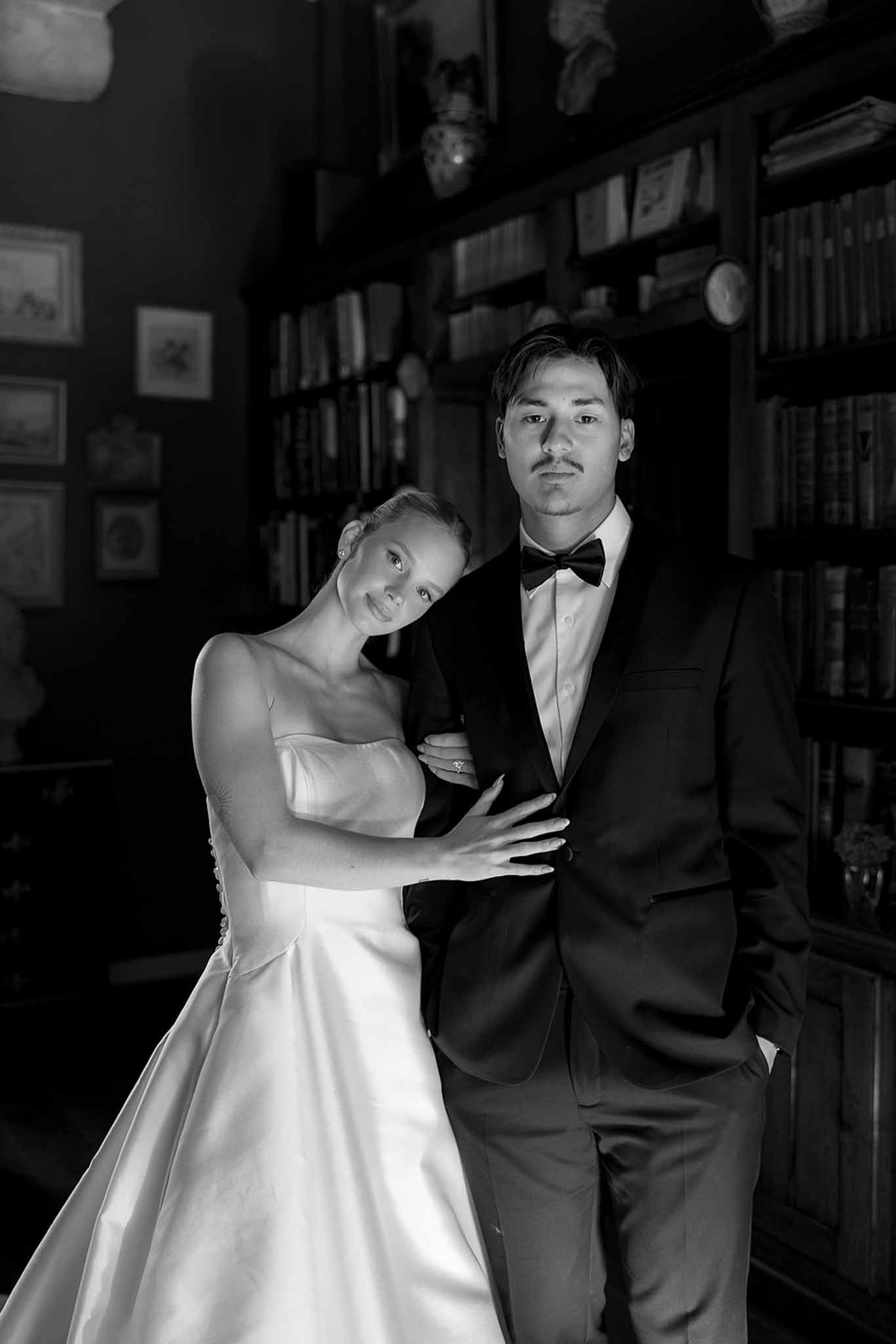 Black and white portrait of bride in strapless ballgown leaning on groom in tuxedo in library setting