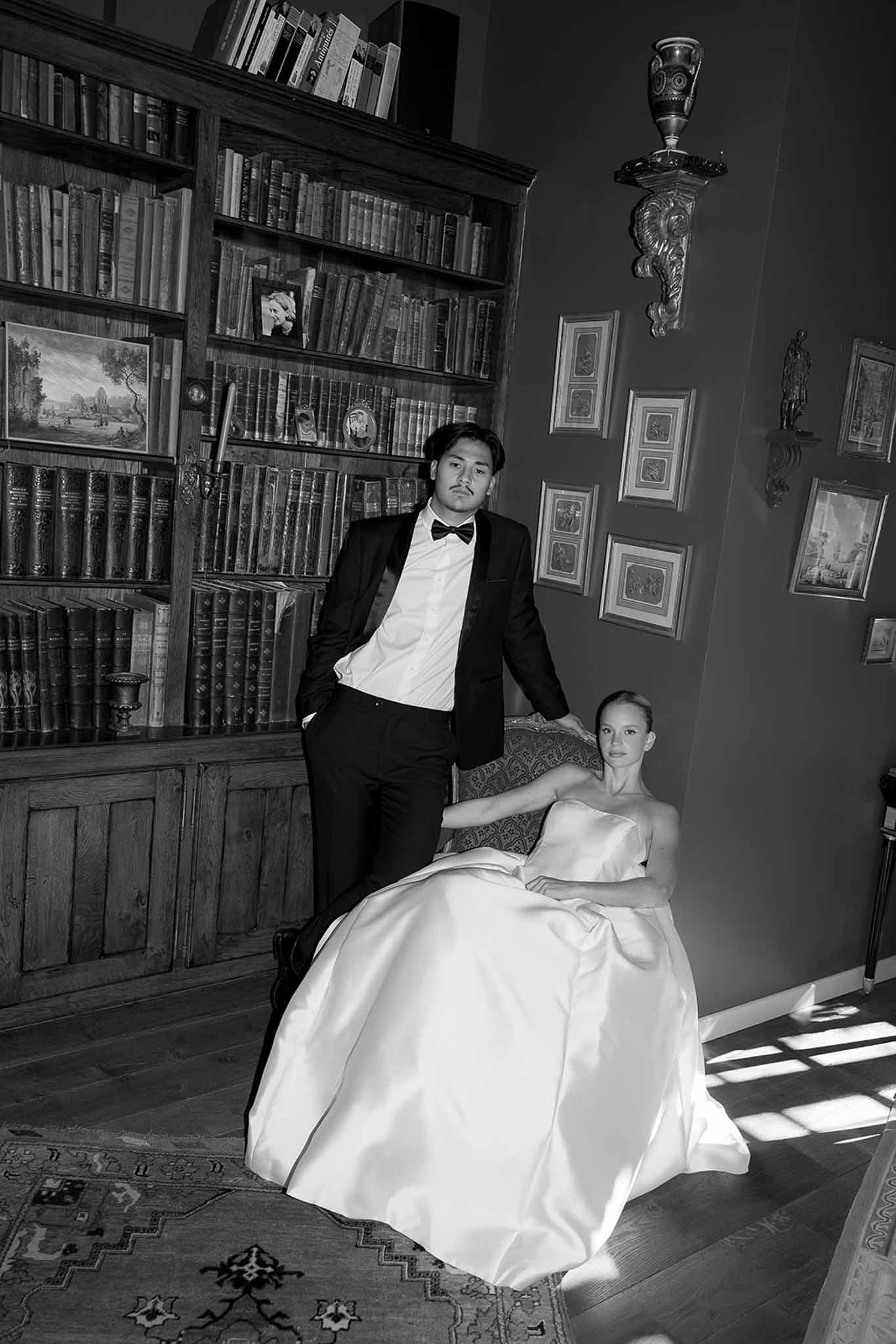Black-and-white portrait of bride seated and groom standing in chateau library with floor-to-ceiling bookshelves