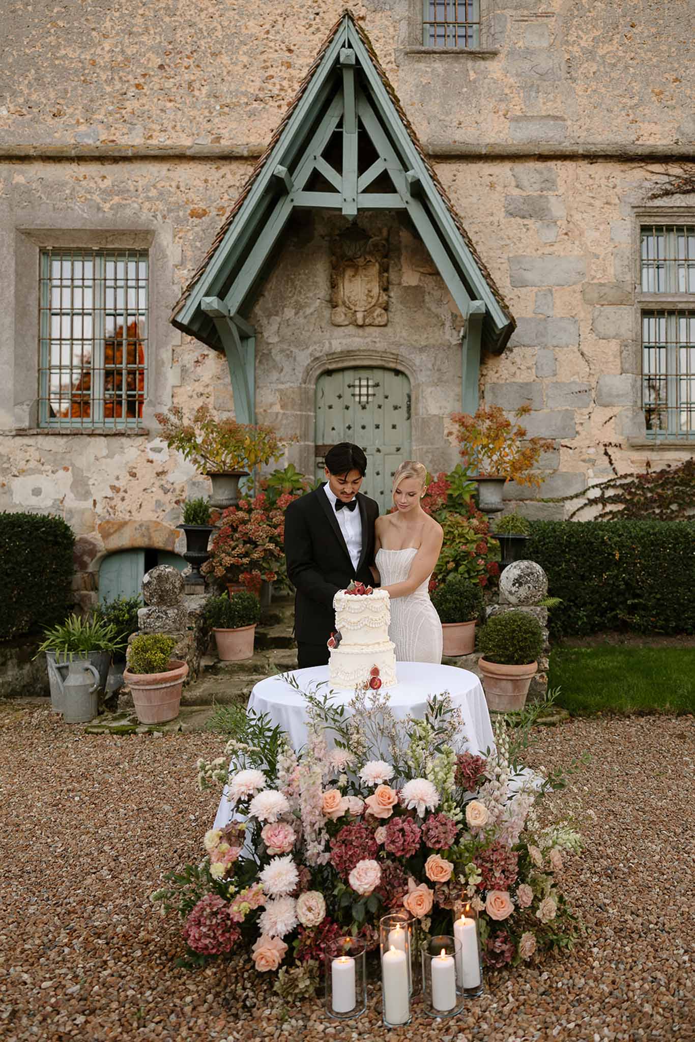 Couple cutting a three-tier white wedding cake surrounded by blush roses and candles at a French manor