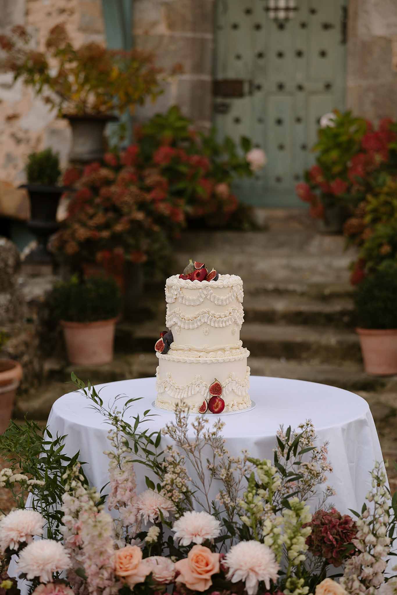 Three-tier buttercream wedding cake with fresh figs and berries surrounded by roses and dahlias outdoors