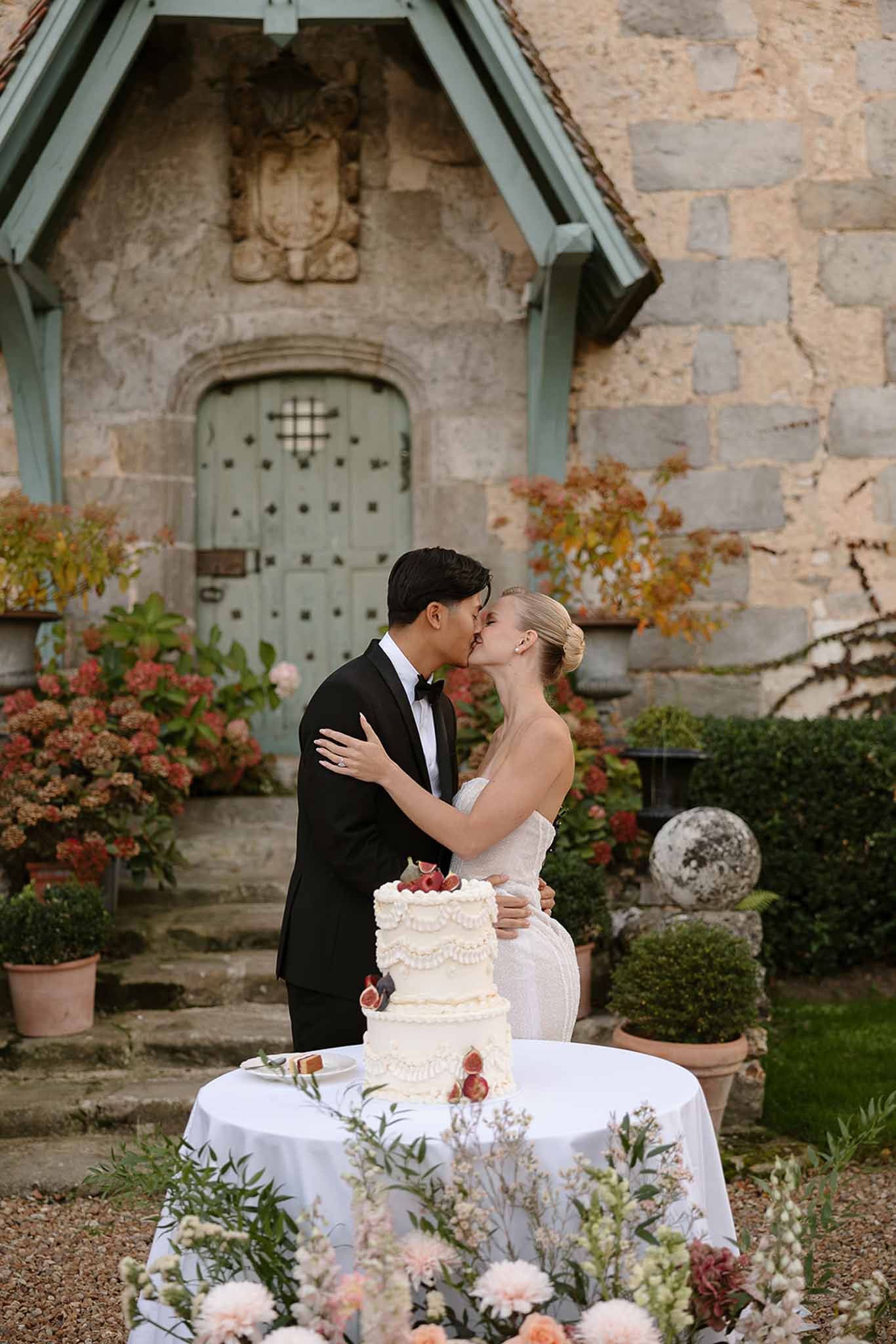 Bride and groom kiss after cutting three-tier ruffled cake with fresh figs at French chateau courtyard