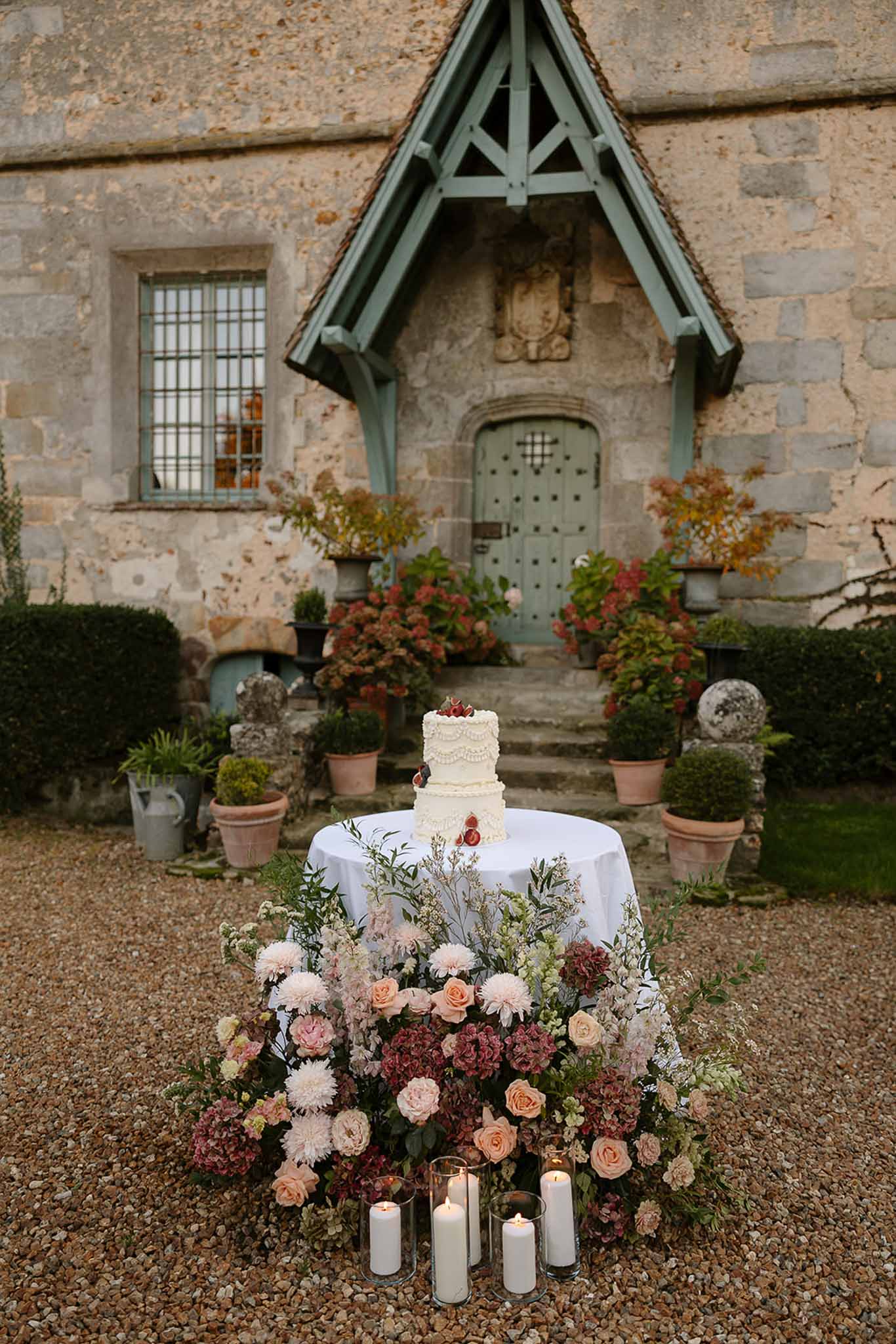 Three-tier fruit-topped cake with blush rose and burgundy hydrangea ground arrangement at stone porch
