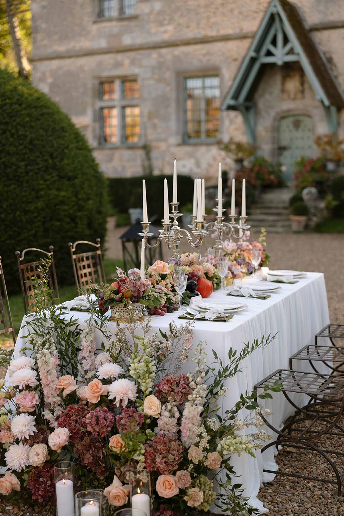 Outdoor reception table with autumn florals, silver candelabras, and green napkins before stone manor