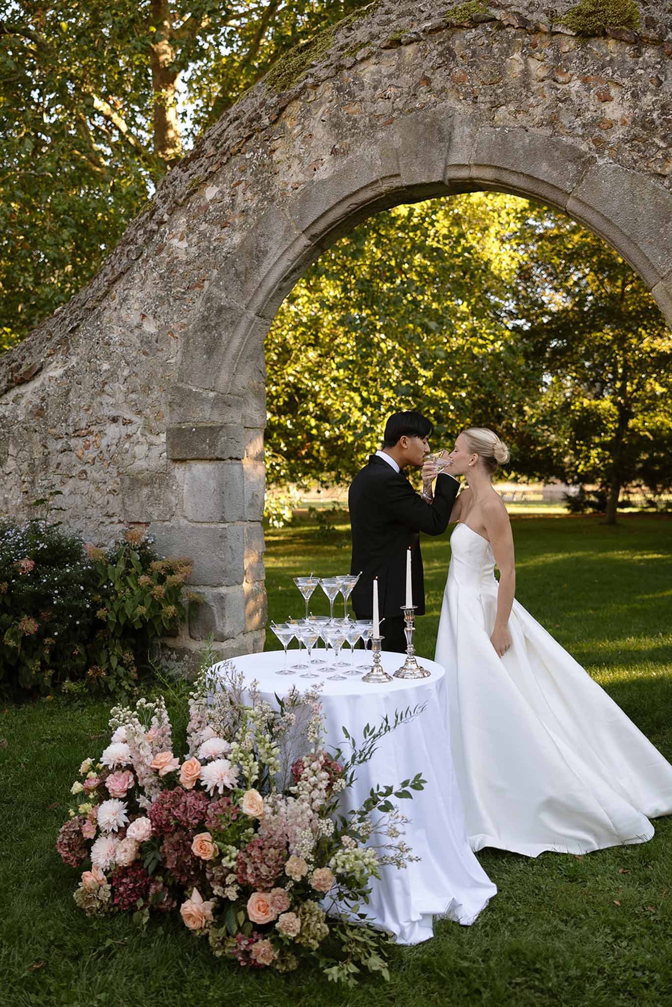 Bride and groom toast champagne coupes beside styled table with glass tower and blush floral arrangement under stone arch
