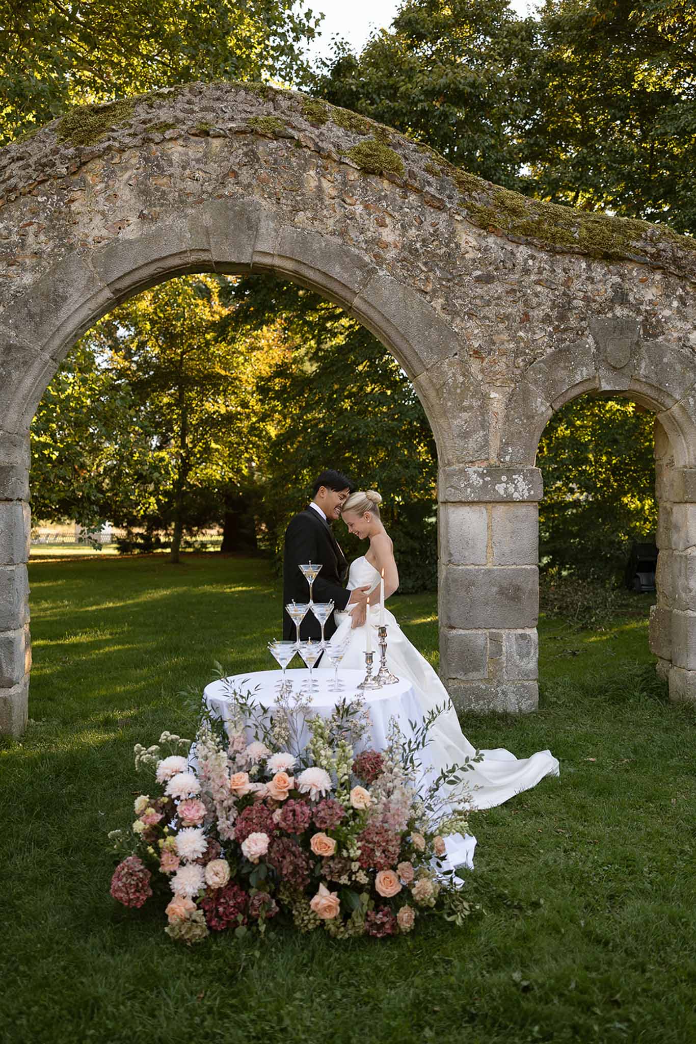 Couple behind martini glass tower under medieval stone arch with blush rose and mauve dahlia arrangement
