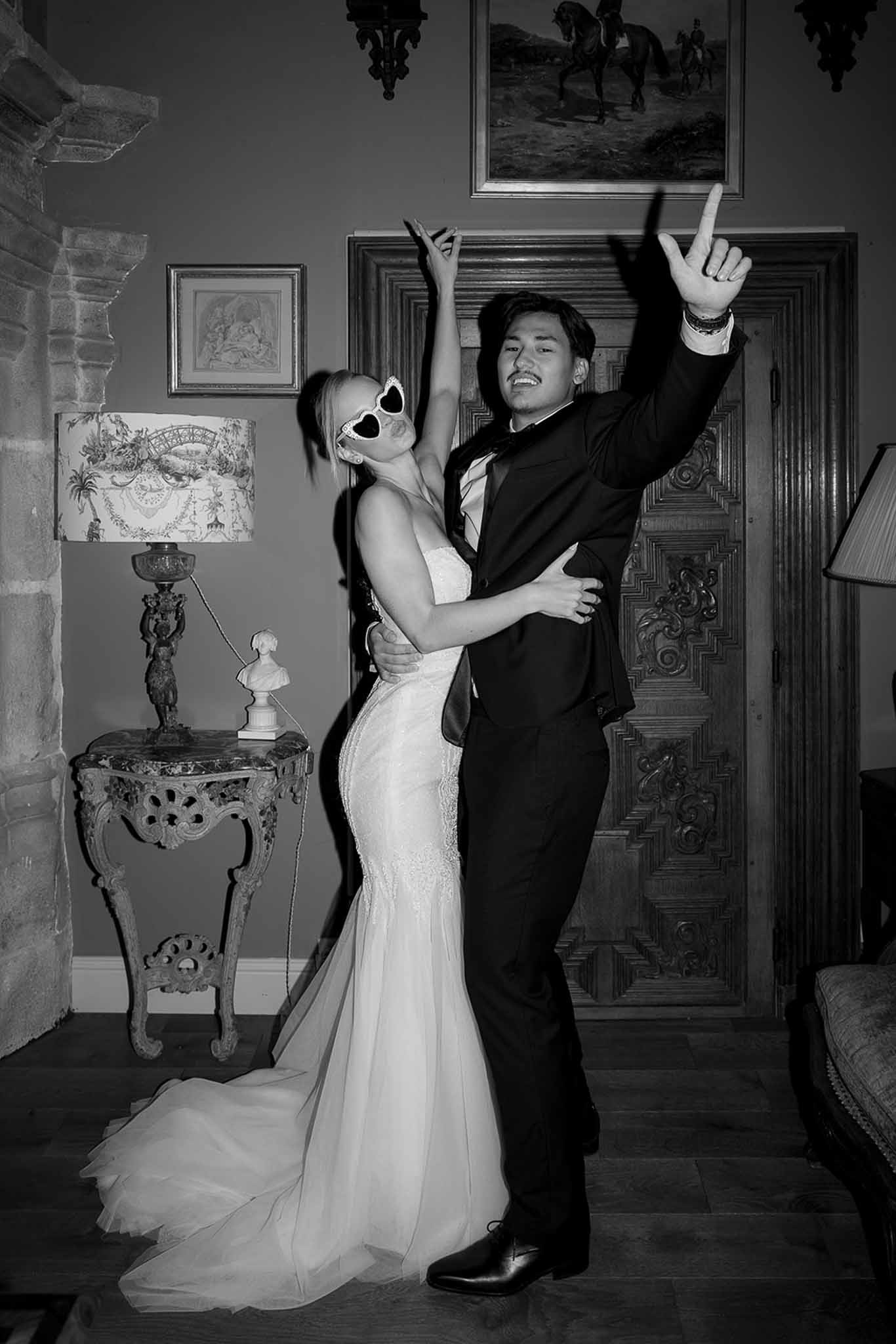 Black and white couple with arms raised celebrating in chateau salon, bride in lace gown with novelty sunglasses