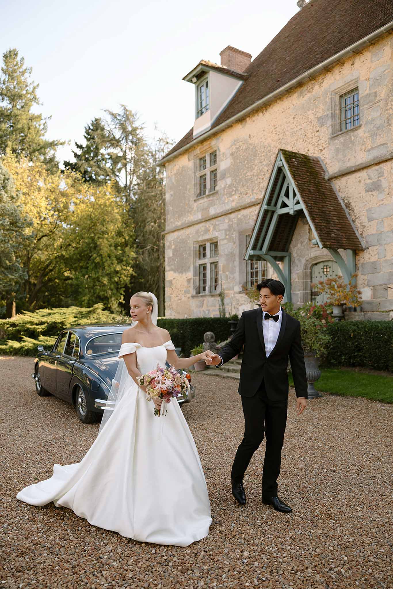 Bride in off-the-shoulder white ball gown and groom in black tuxedo walking hand in hand with vintage navy car behind
