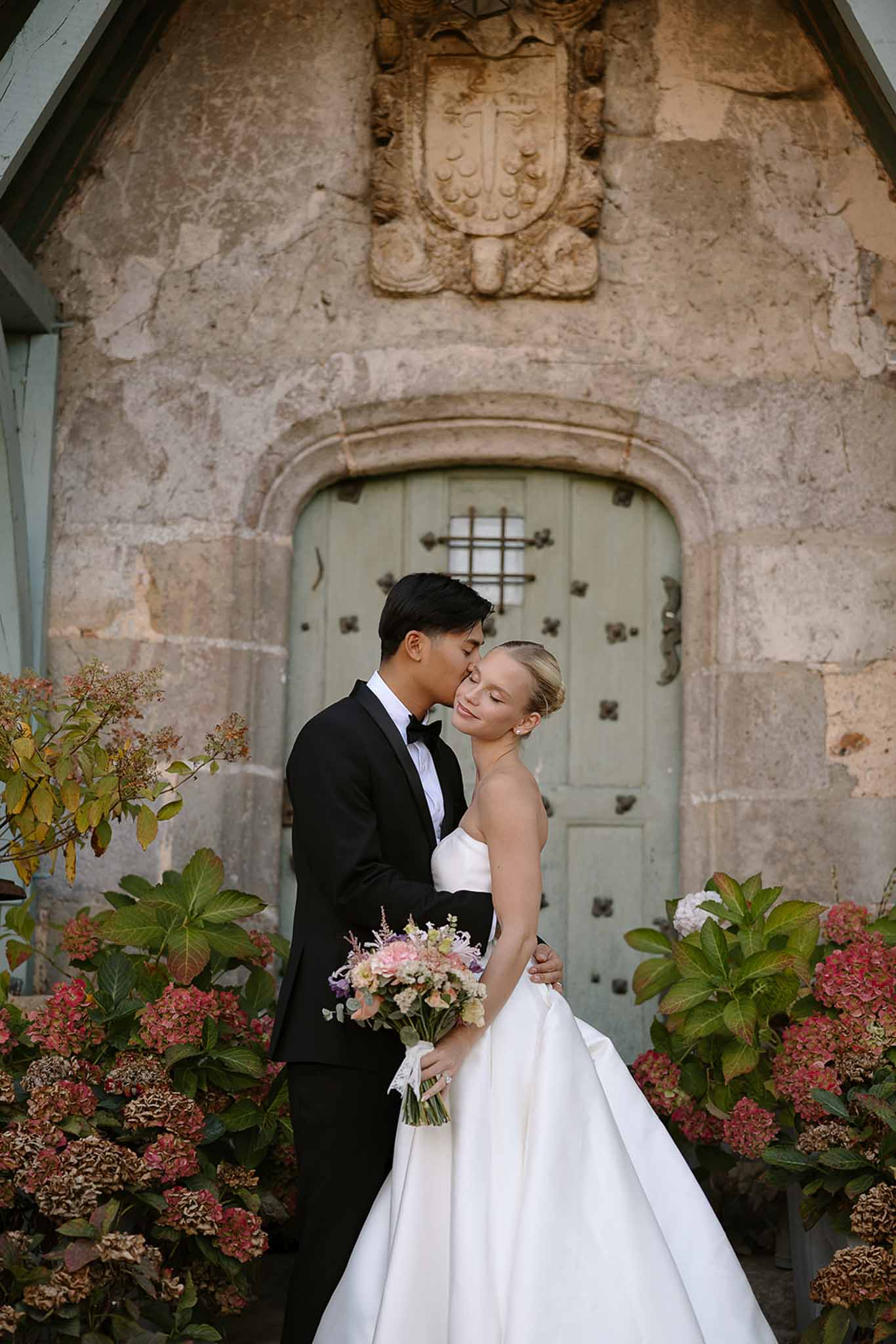 Groom kissing bride on temple beside sage green door with coat of arms, holding blush and lavender garden bouquet