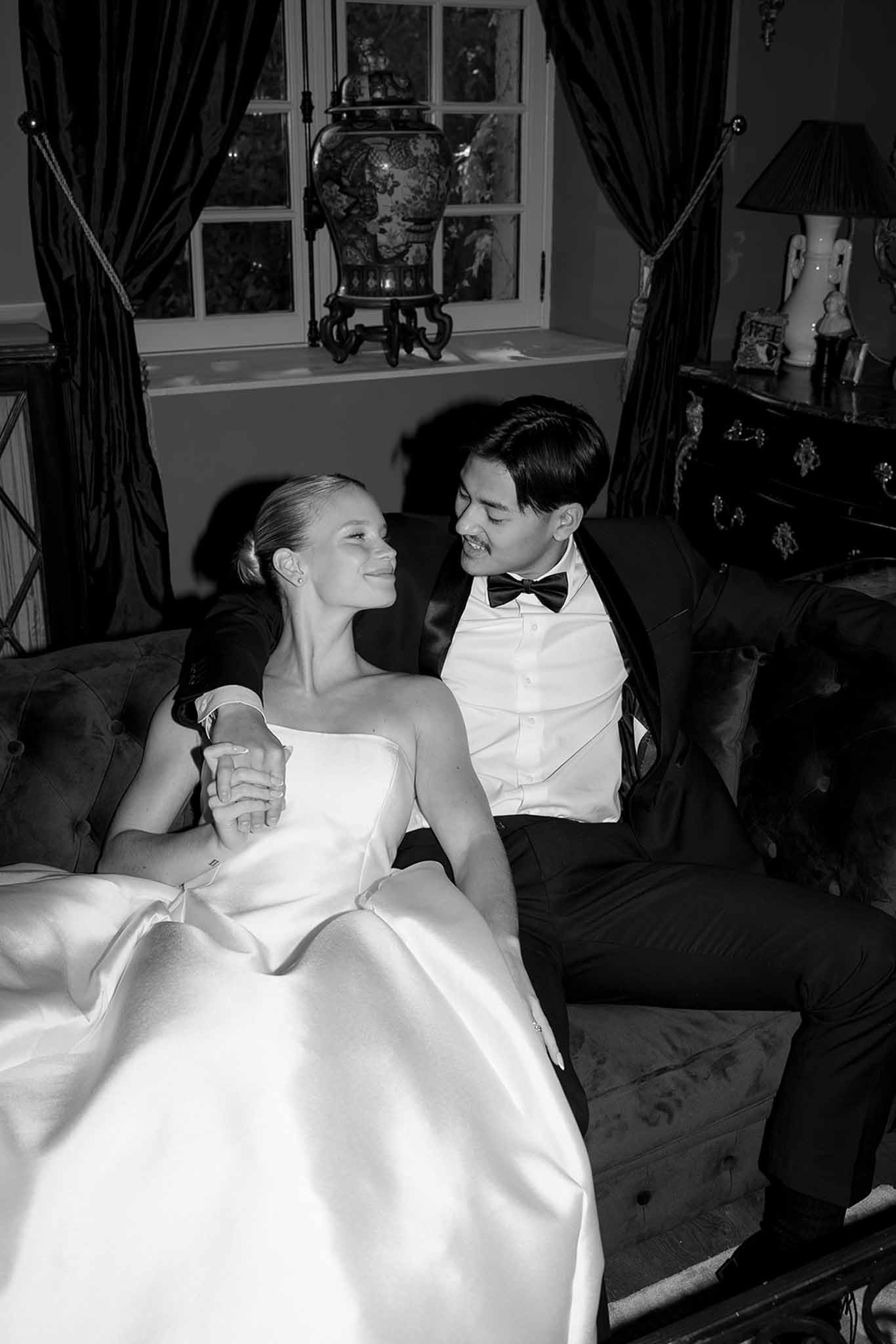 Black and white portrait of bride and groom seated on tufted sofa holding hands in ornate chateau interior