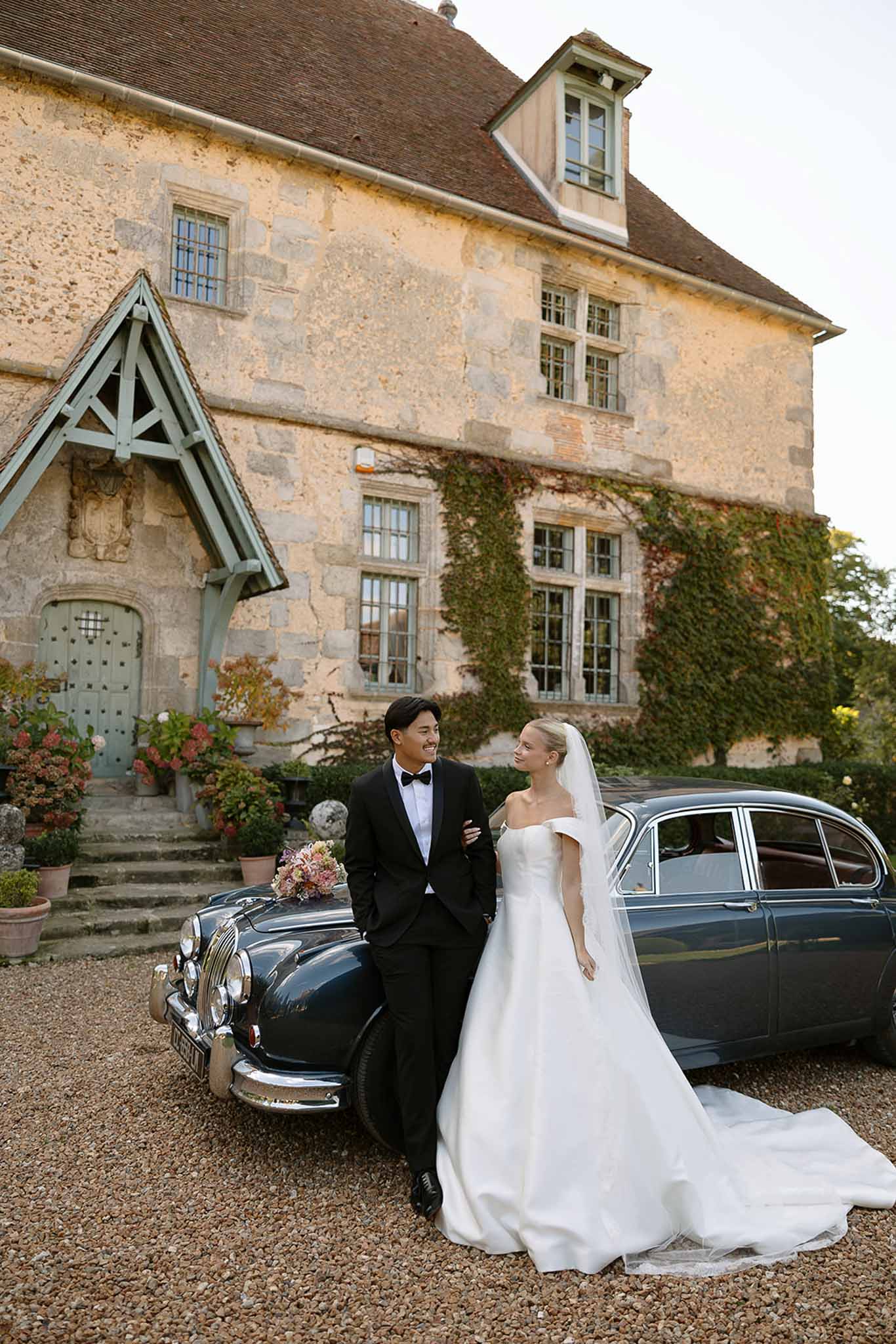 Bride and groom lean against vintage Jaguar on gravel drive in front of ivy-clad French manor house
