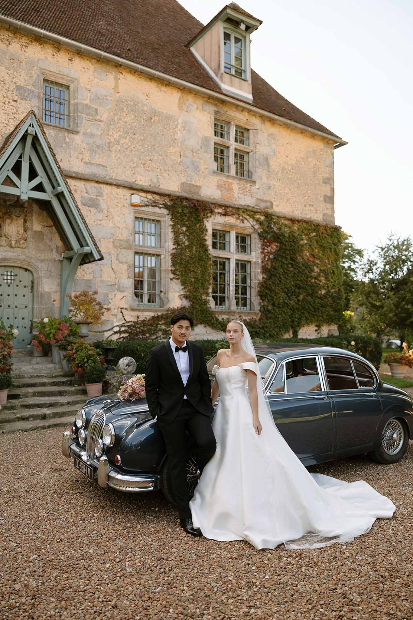 Bride in ballgown with veil and groom in tuxedo lean against vintage Jaguar before stone manor