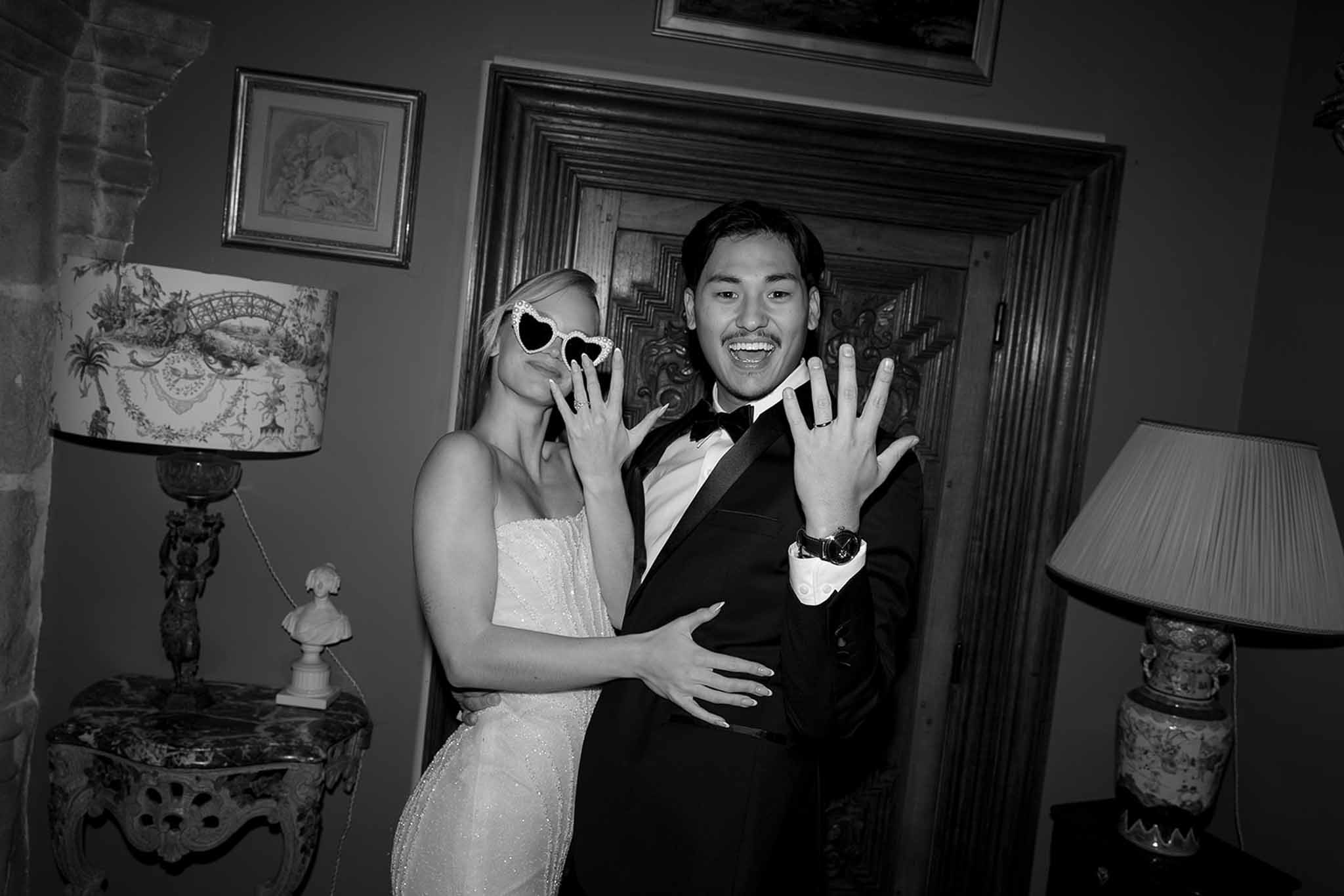 Black-and-white portrait of bride and groom showing wedding rings in an ornate chateau interior with carved doors