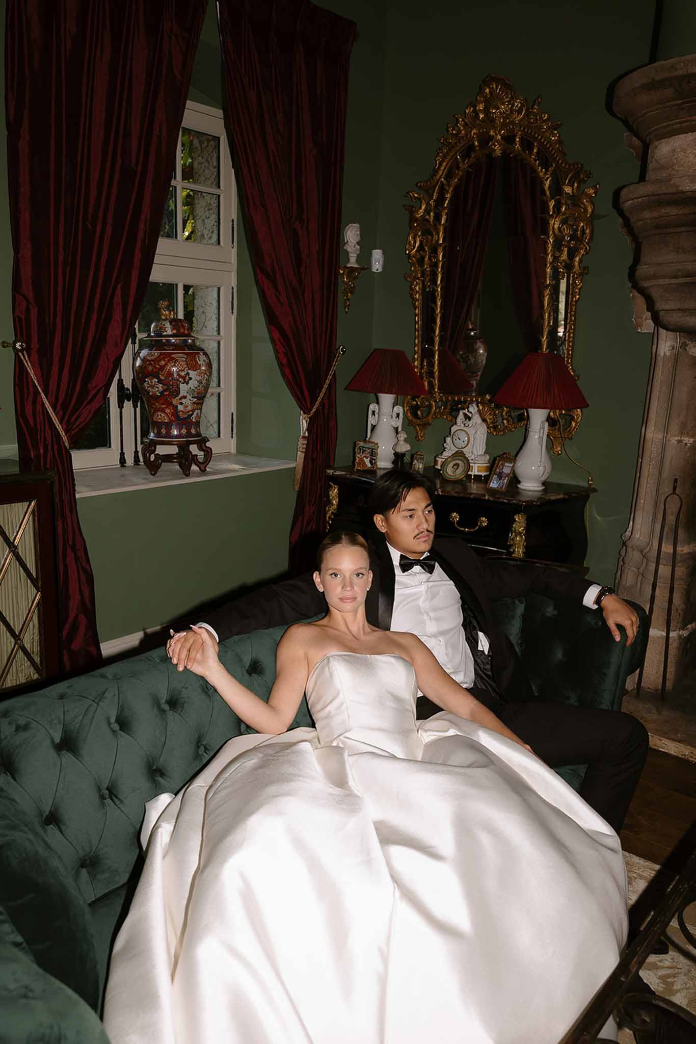 Bride in strapless ivory ball gown and groom in black tuxedo seated on green velvet sofa in chateau salon