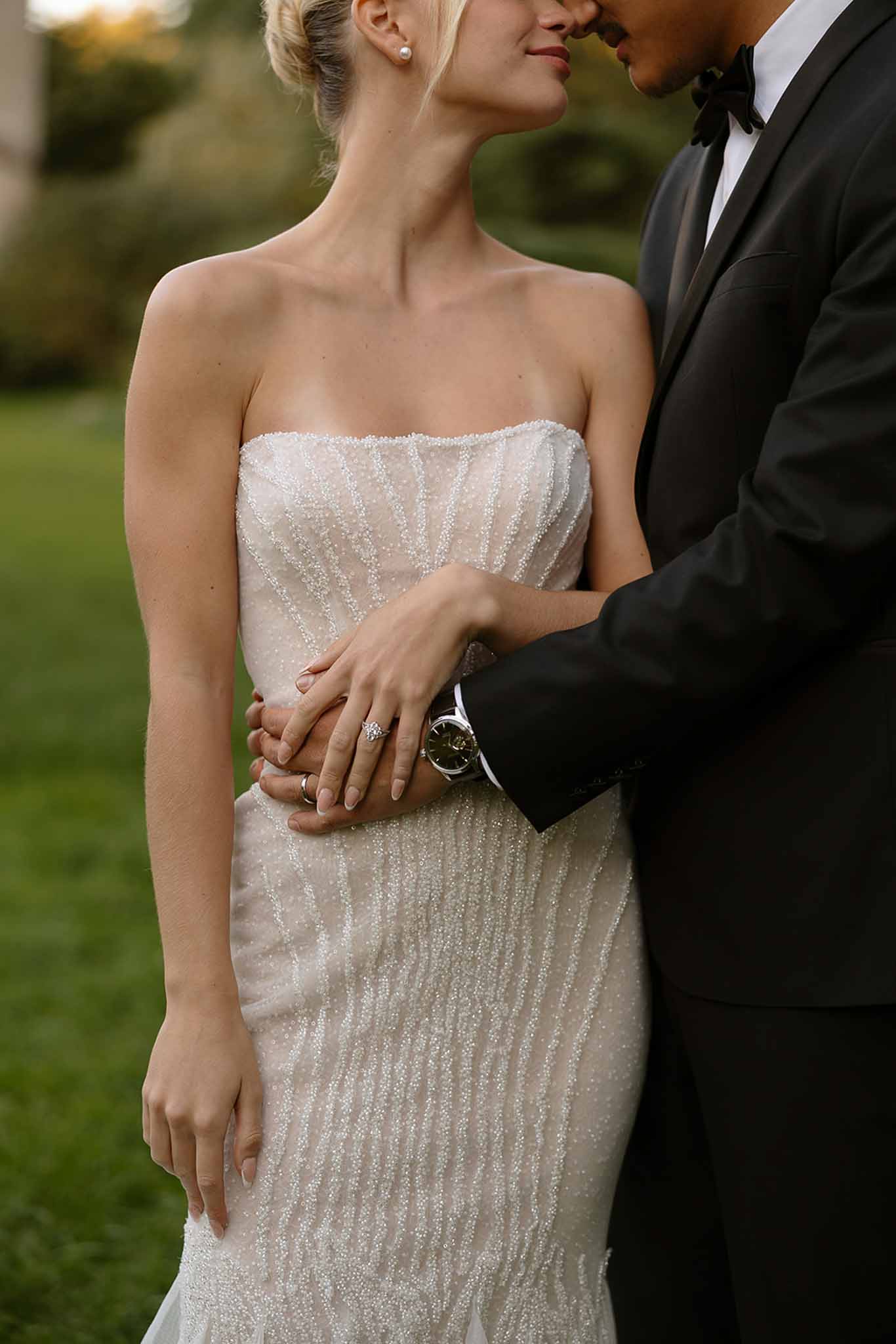 Close-up of bride in beaded gown and groom in tuxedo embracing with interlaced hands and rings