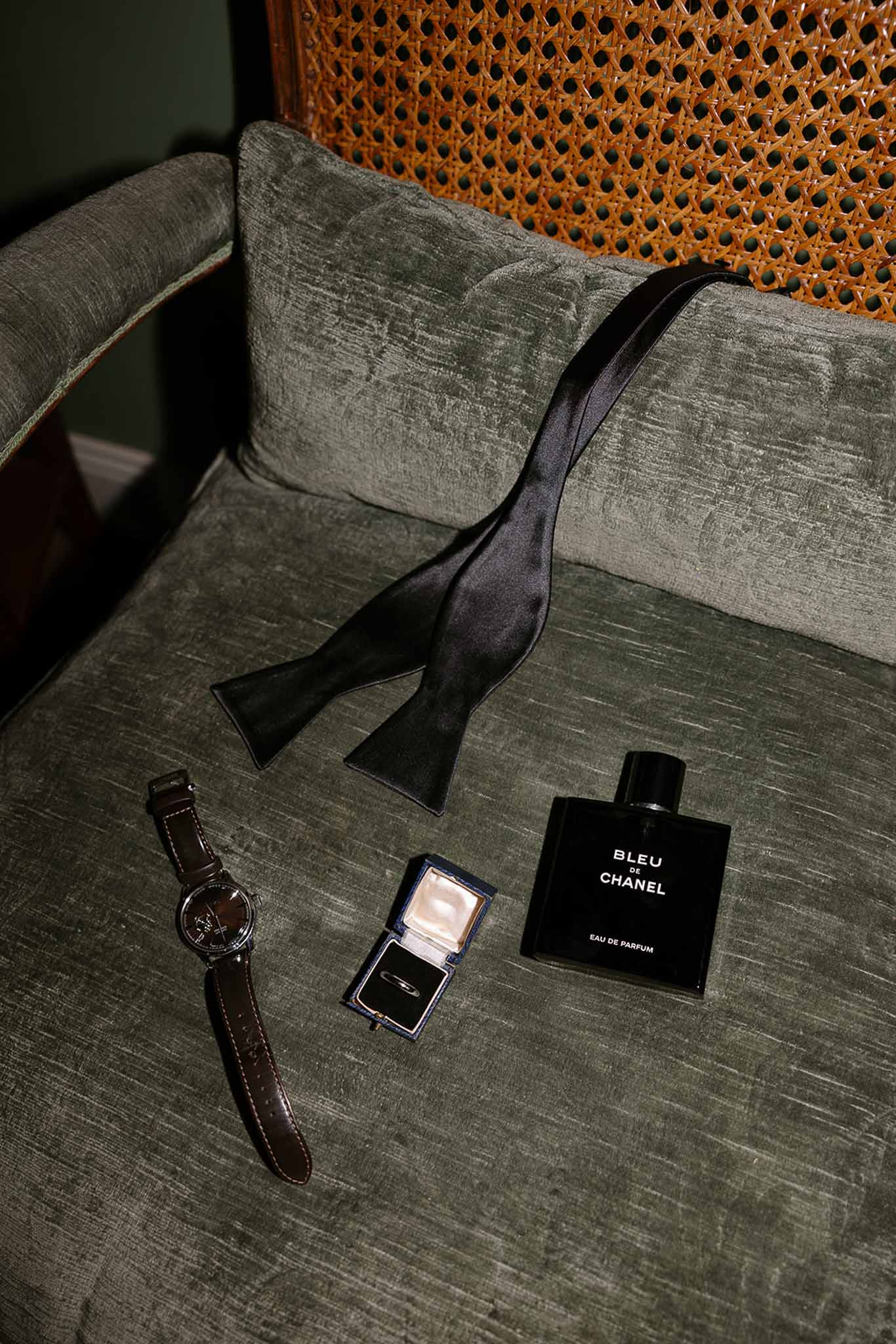 Groom flat lay on sage velvet chair with bow tie, watch, gold wedding band and Bleu de Chanel cologne