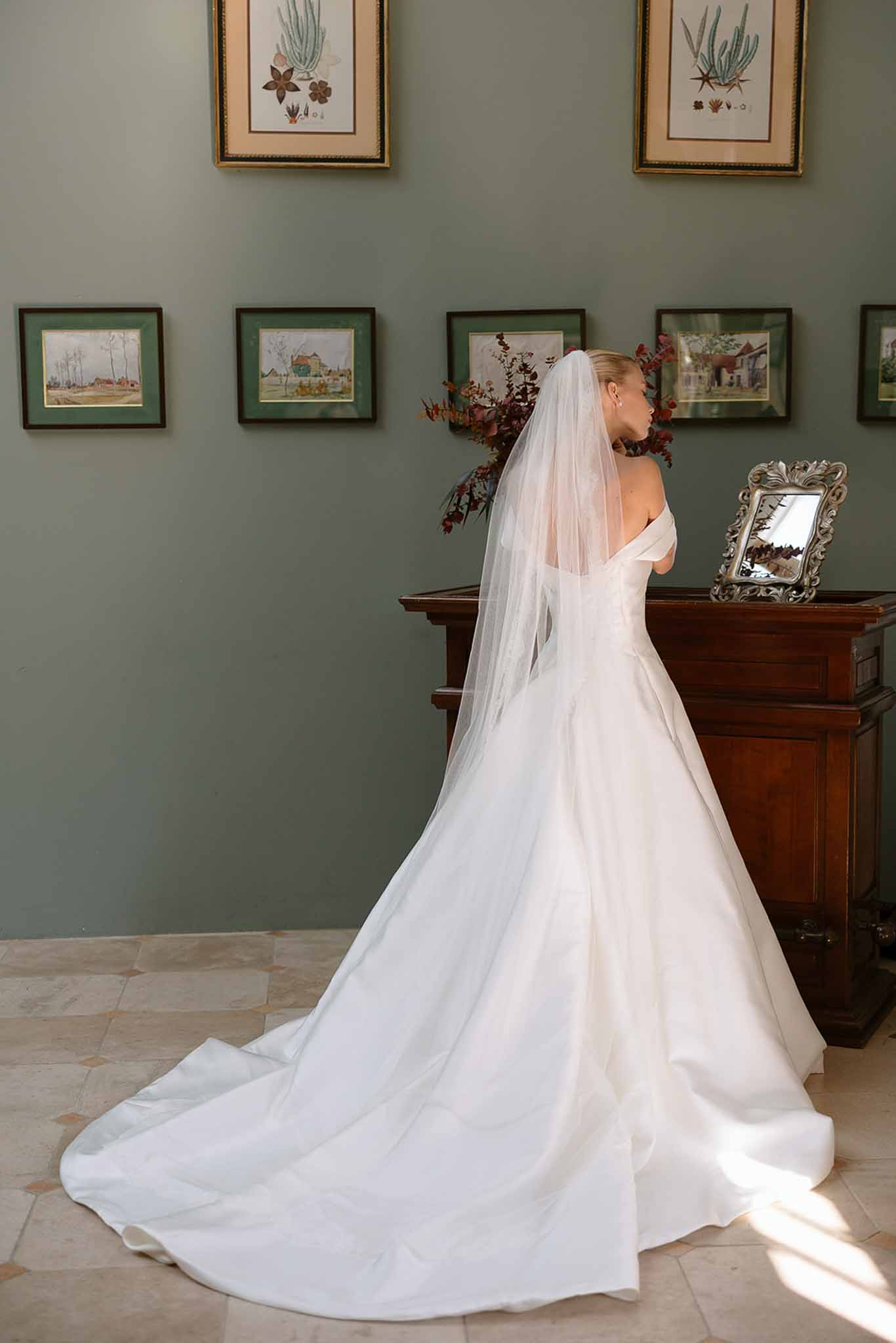 Bride from behind in ivory ballgown with cathedral veil beside burgundy bouquet and botanical gallery wall