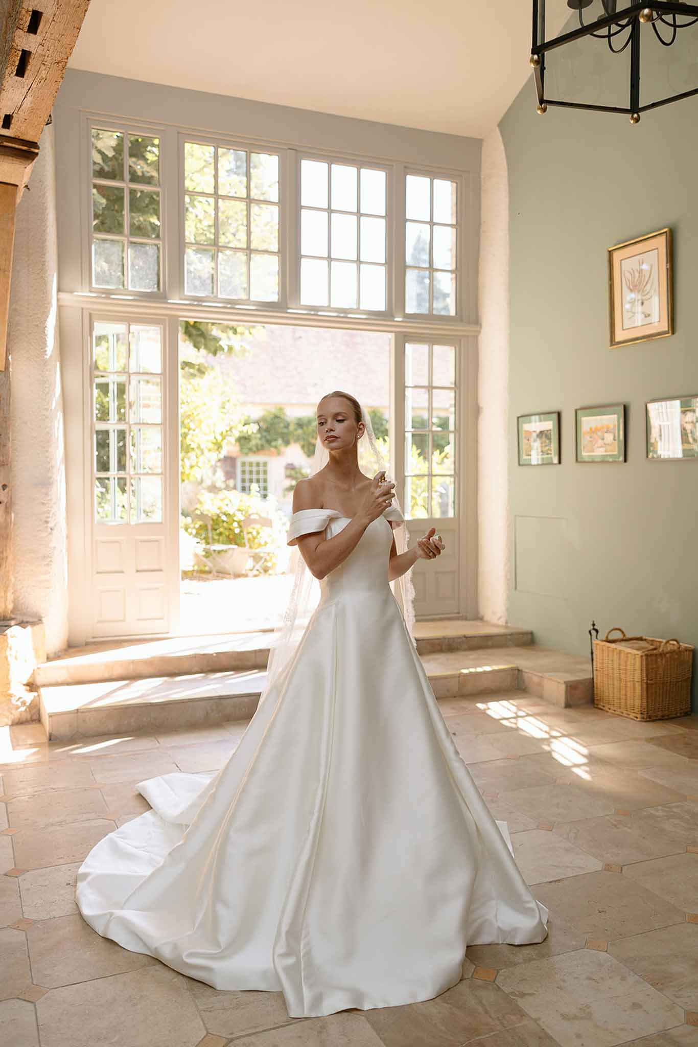 Bride in off-shoulder satin ball gown holding veil in sage green chateau room with natural light