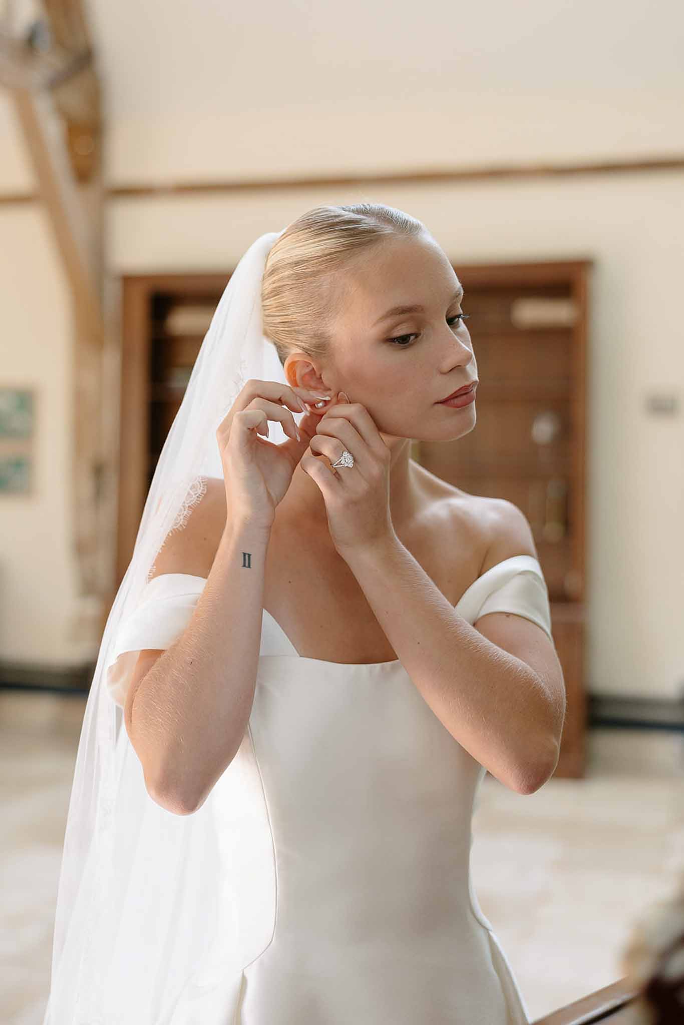 Bride fastens earring in off-shoulder satin gown with lace-trimmed veil over sleek low bun close-up