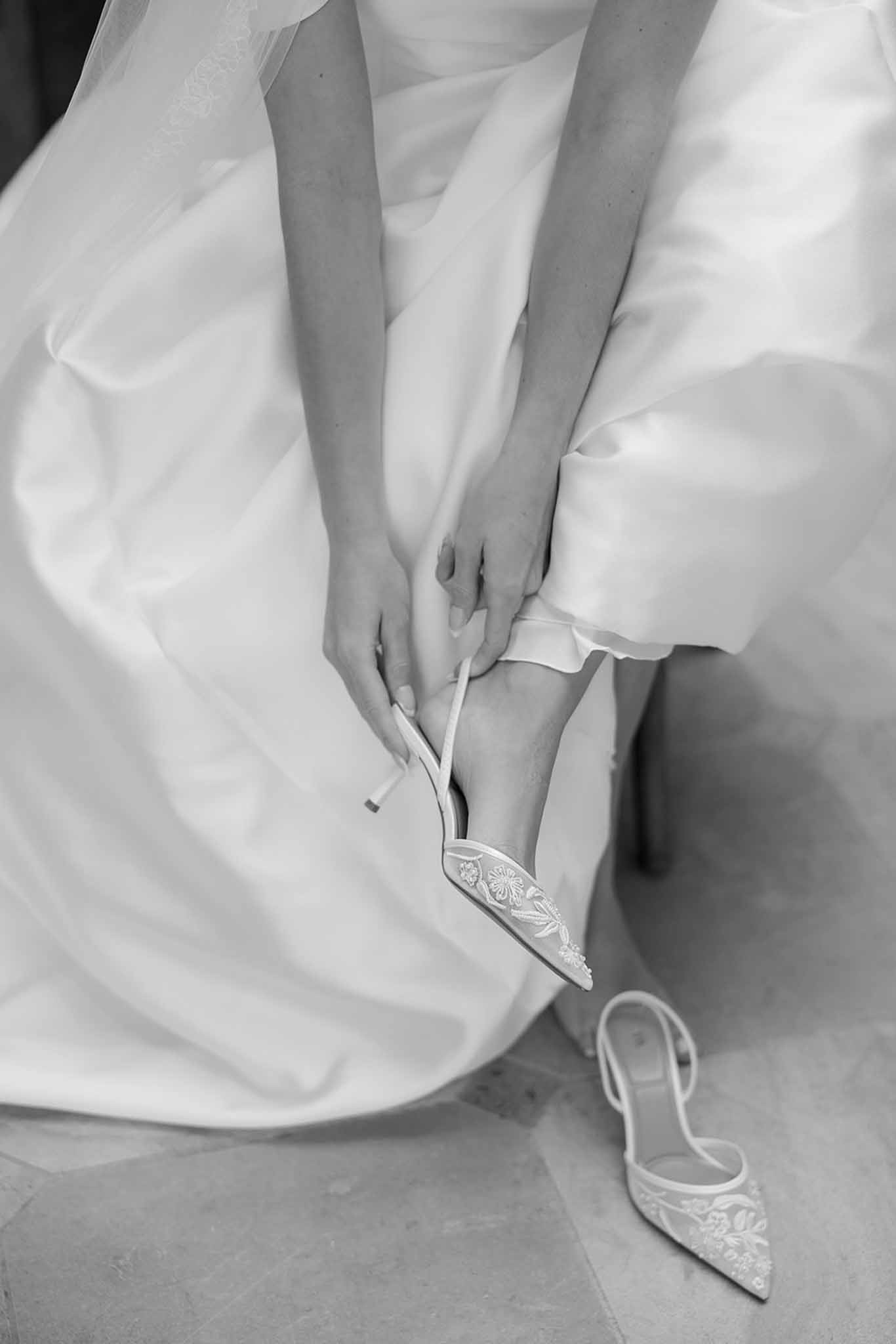 Black and white of bride fastening embroidered slingback heel with satin gown pooling around her on tiled floor