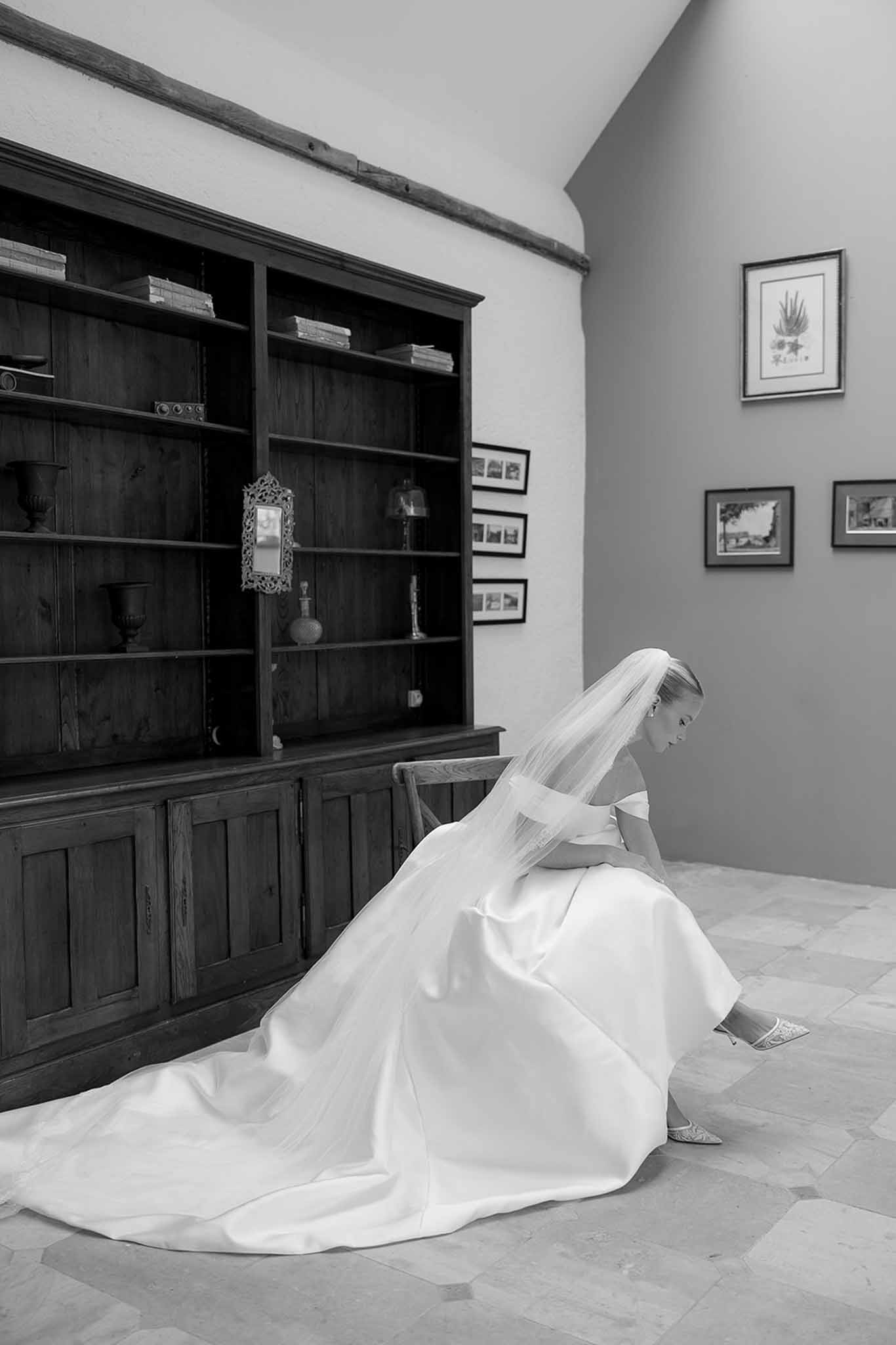 Black and white bride adjusting heel in ballgown with cathedral veil beside dark wood bookcase and framed artworks