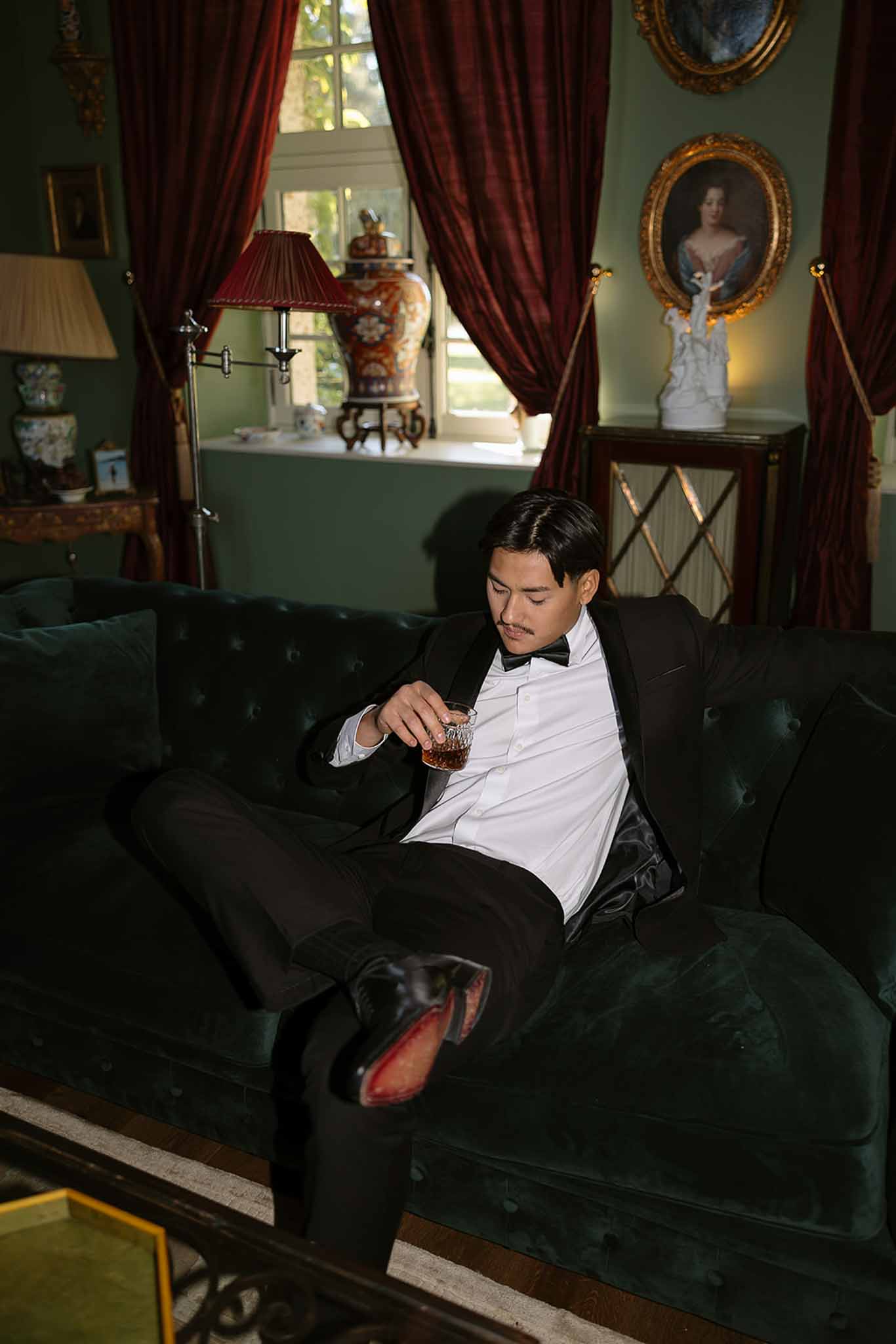 Groom in black tuxedo with bourbon glass on green velvet sofa in burgundy-curtained chateau salon