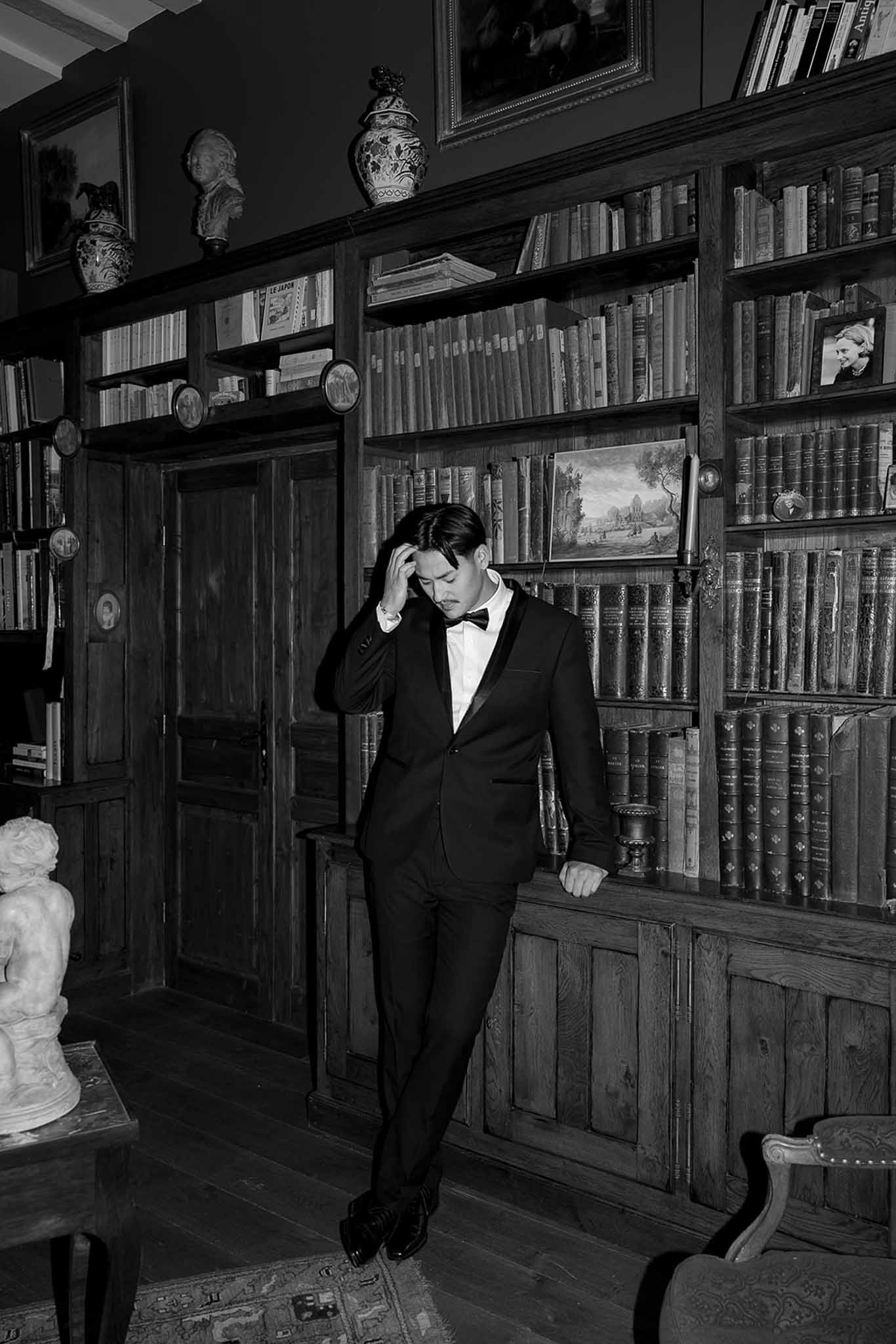 Black and white portrait of groom in tuxedo standing in chateau library with antique bookshelves