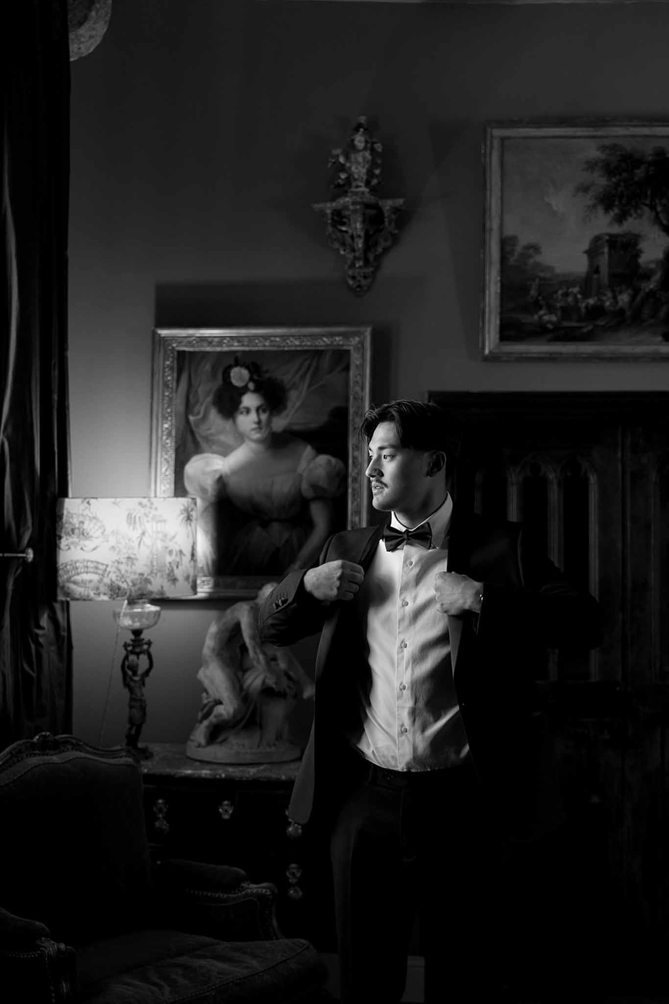 Black and white groom adjusting jacket in chateau room with gilt portrait and antique lamp