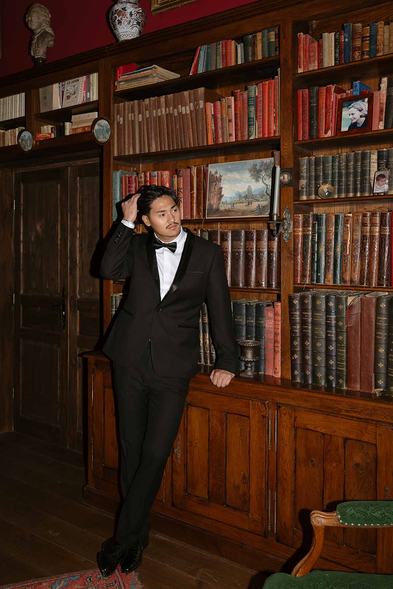 Groom in tuxedo leaning against floor-to-ceiling walnut bookshelves in chateau library with red walls