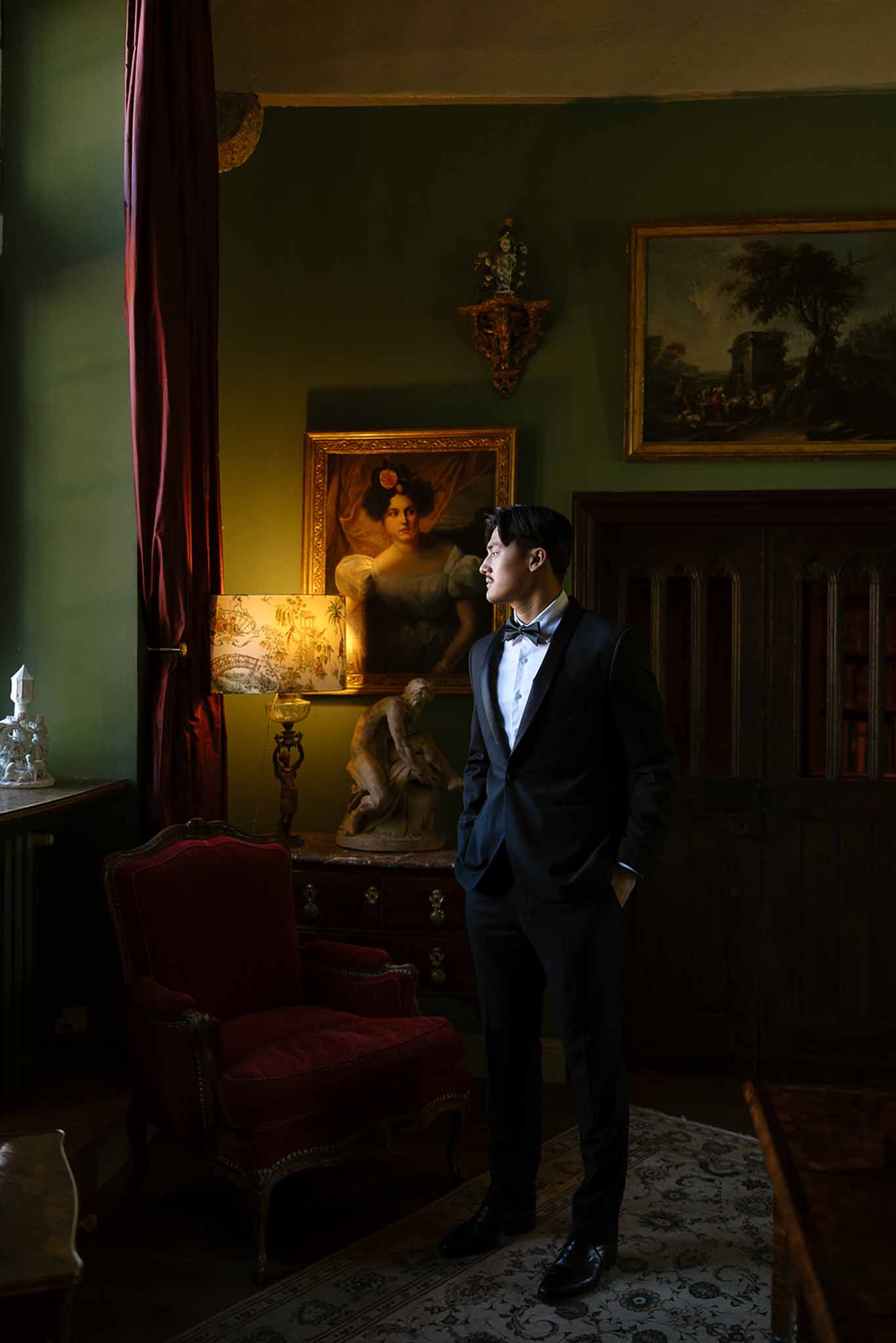Groom in tuxedo stands in olive-green salon with gold-framed paintings, red velvet chair, and warm lamp