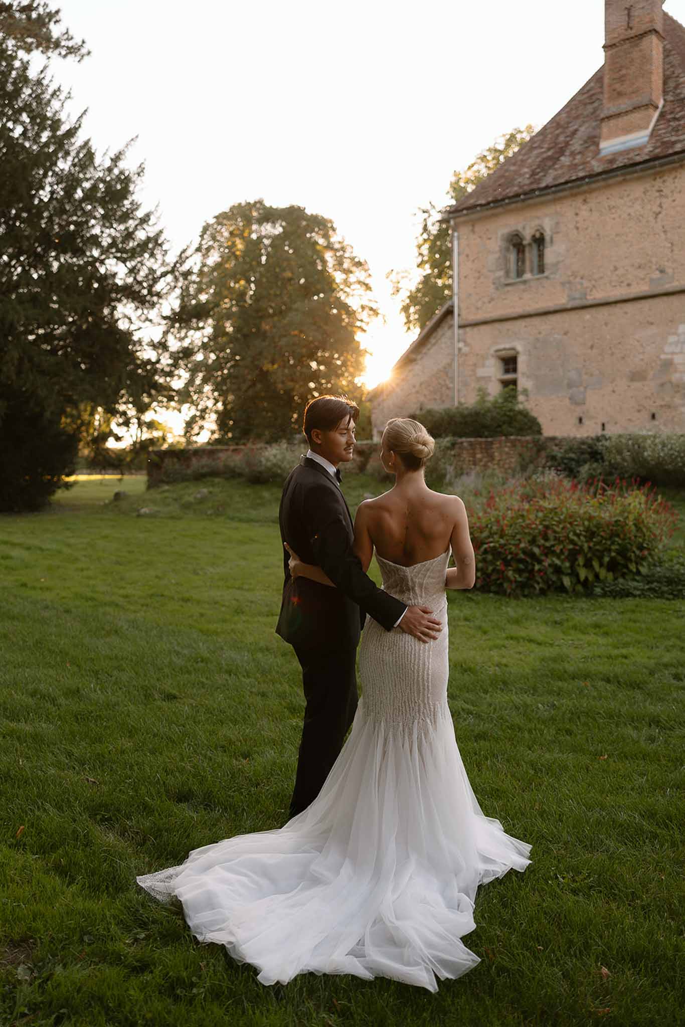 Bride in beaded corset gown and groom in navy suit photographed from behind at golden-hour manor