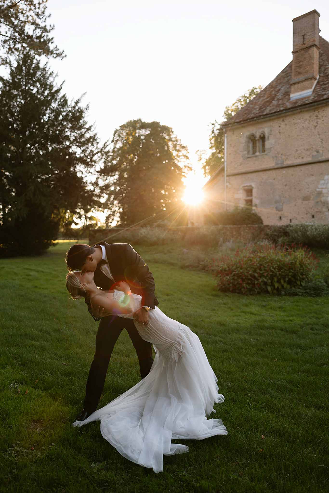Groom dips bride for a kiss at golden hour on chateau lawn with sun flare through trees
