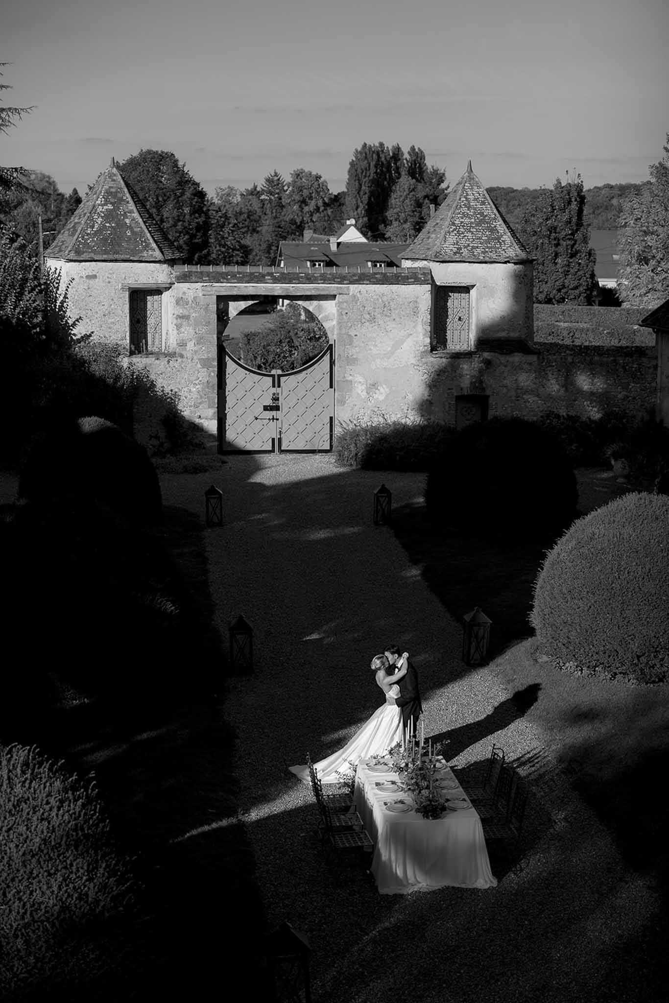 Black and white aerial couple kissing beside reception table in formal garden with turreted gatehouse