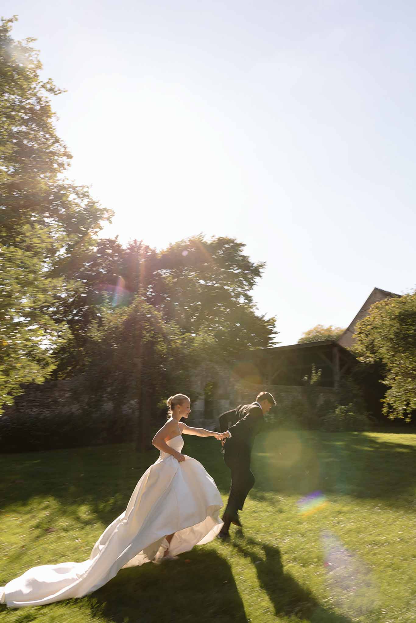 Bride and groom running across estate lawn with cathedral train sweeping grass in golden-hour backlight